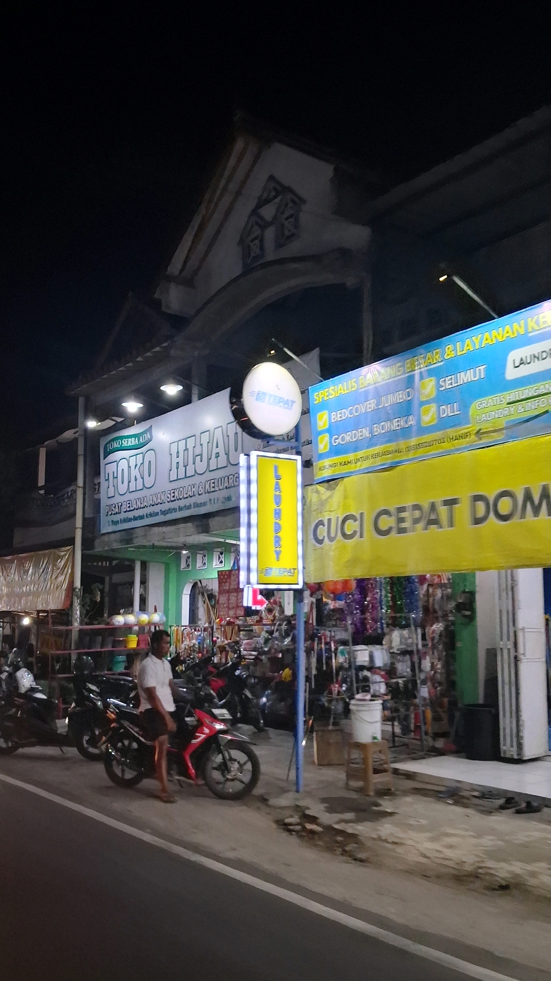 In the city of Berbah, Indonesia, an outdoor scene at night features a street with several motorcycles parked on the side, with one red motorcycle in the foreground with a man in a white shirt and dark shorts standing next to it. Behind the motorcycles, there are various shops and stalls illuminated by bright signs. One prominent sign reads "TOKO HIJAU" and another announces "LAUNDRY" in yellow lettering with "TEPAT" logo. A larger yellow banner displays "CUCI CEPAT DOM". The shops appear to sell a variety of goods, including household items and clothing. The overall atmosphere suggests a bustling commercial area at night.