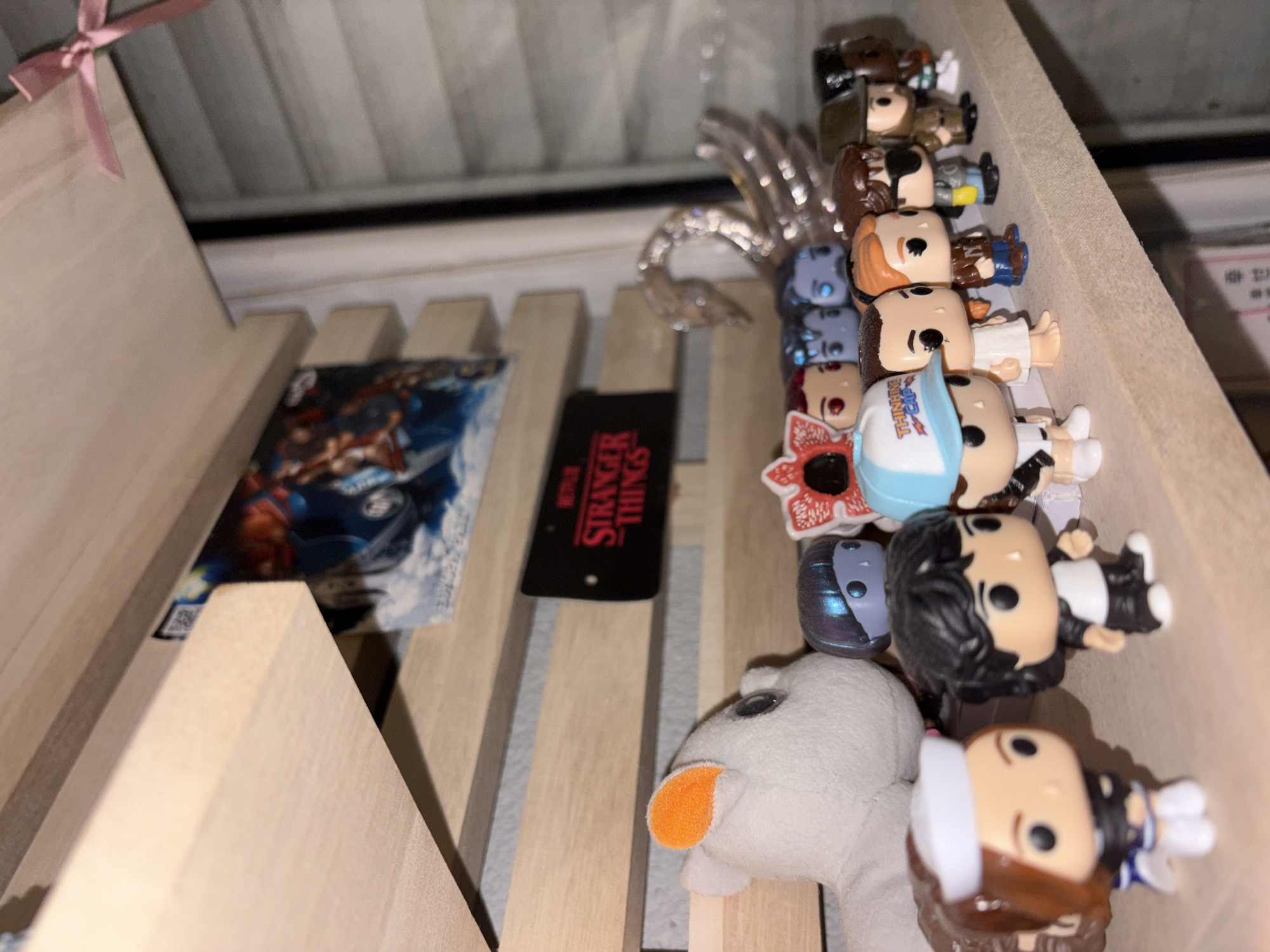 The image shows a collection of items within a wooden box or tray, possibly a storage container or display case. The items include:
* Several Funko Pop figurines, apparently from the "Stranger Things" series, are arranged in a row.
* A "Stranger Things" branded item, possibly a tag or label, is visible.
* A small plush toy, possibly a sheep, is among the items.
* A clear, possibly glass, decorative piece with an ornate, curved design is present.
* A trading card or collectible card with a blue and white design is also present.
There is no information to confirm the location is Panagyurishte, Bulgaria.