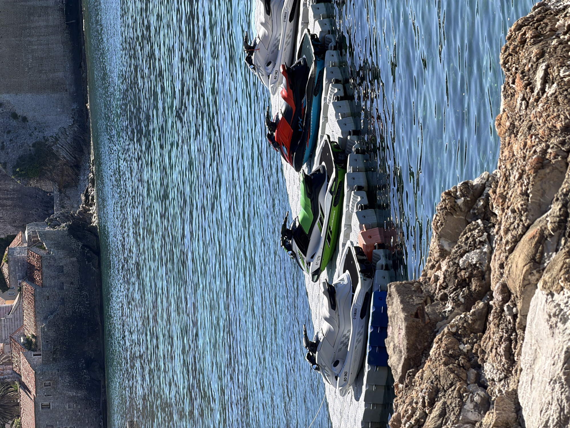 The image shows a row of jet skis docked on a floating platform next to a rocky shoreline in Budva, Montenegro. The water is calm and blue. In the distance, the old town of Budva is visible with its stone buildings and terracotta roofs. The jet skis are of different colors, including white, green, red, and blue. The rocky shoreline is rough and uneven.