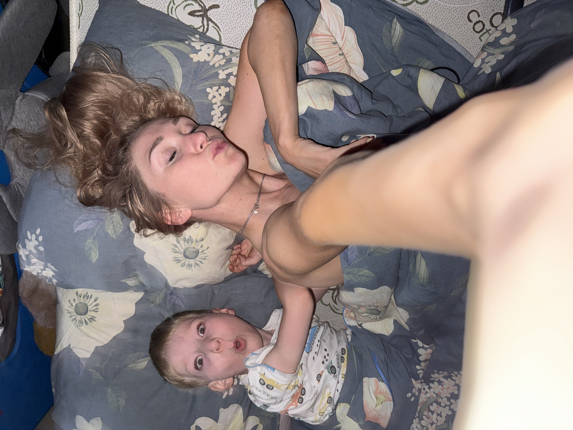 A woman and a young child are lying on a bed covered with a floral-patterned bedspread. The woman is on top, with her head at the upper left of the frame. She has light brown hair, pulled back and styled, and is making a kissy face toward the camera. She is wearing a silver necklace. The child, who appears to be a young boy with blond hair, is positioned below her, with his head at the lower left of the frame. He is wearing a white shirt with vehicles printed on it and is looking at the camera with his mouth open in a surprised expression. The bedsheets feature a blue-gray background with large white and gray floral prints. The environment around the bed is partially visible. The location is Chaikovsky, Russia.