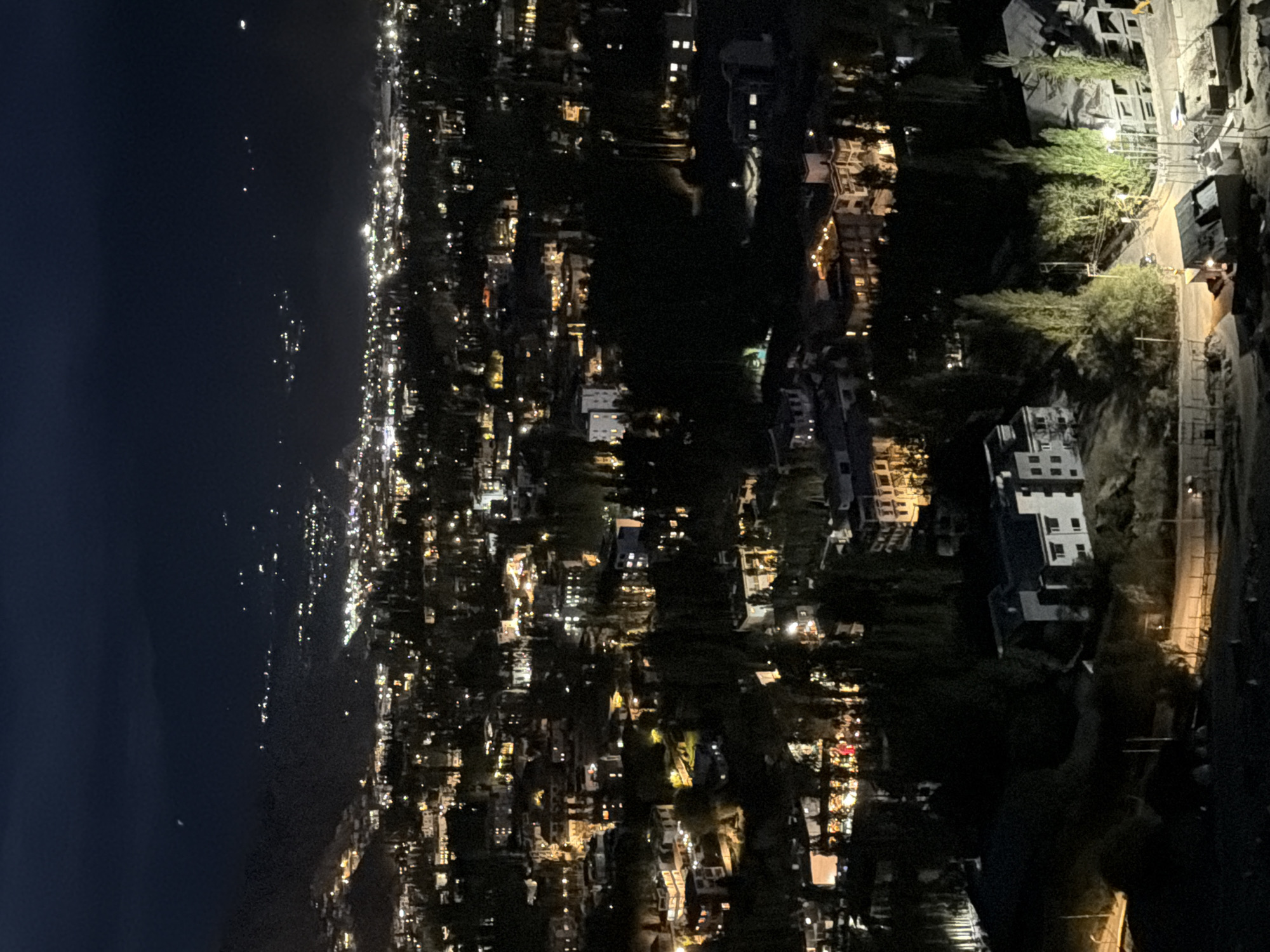 This is a nighttime aerial view of the city of Leh, India. The image captures a sprawling urban landscape illuminated by numerous building lights. In the foreground, closer buildings are visible, some with multiple stories and lit windows. Further into the background, the city extends across a valley, with lights receding into the distance and forming a dense pattern. The surrounding terrain appears mountainous and dark, with only faint indications of distant lights suggesting hills or other settlements on the periphery. The overall scene depicts a densely populated urban area at night, showcasing the artificial light emanating from buildings against the dark natural backdrop.