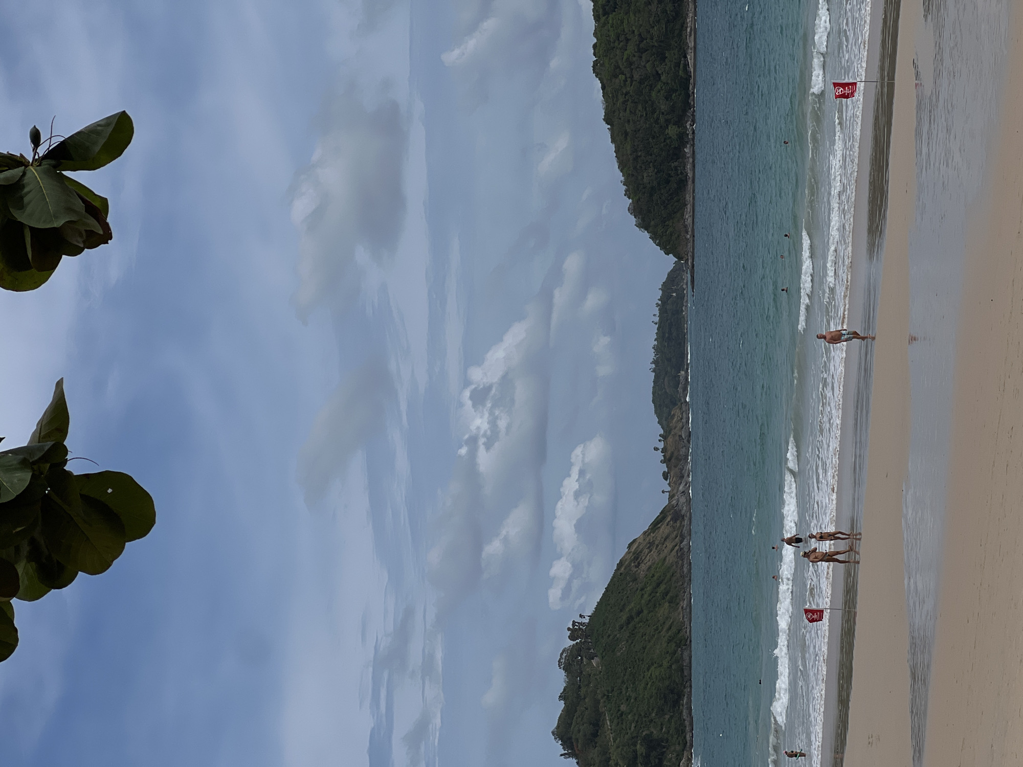 The image depicts a beach scene. A section of coastline is visible, with green, foliage-covered land masses on either side of a sandy beach. The ocean has varying shades of blue and white, indicating waves. Several people are visible on the beach and in the water. Two red flags are positioned on the beach, possibly serving as warnings or markers. The sky is filled with clouds. The location is in the city of Unknown, Unknown.