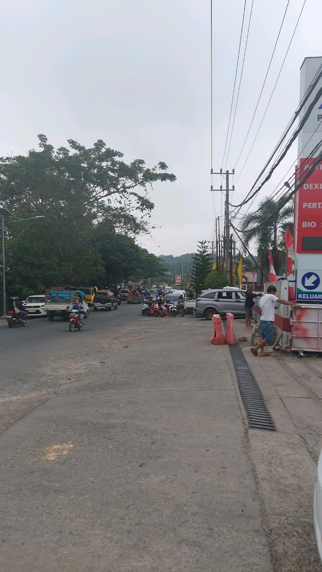 The image depicts a street scene in Tanjung Redeb, Indonesia, with a moderate amount of traffic. In the foreground, a paved road runs diagonally from the bottom left to the center. To the right, there is a white building with a red sign that reads "PERTAMINA" and "KELUAR" in Indonesian, indicating a gas station. Several orange traffic cones are placed on the ground near the entrance to the gas station.
On the road, there are various vehicles including cars, trucks, and motorcycles. A silver SUV is parked near the gas station. A group of motorcycles is also parked on the side of the road. Several people are visible, including individuals standing near the gas station and people riding motorcycles. A man in a white t-shirt and blue shorts is standing near the gas station entrance, looking towards the road.
In the background, there are trees, power lines, and utility poles. A hill is visible in the distance. The sky is overcast. The overall impression is a busy street in a tropical or subtropical urban environment.
