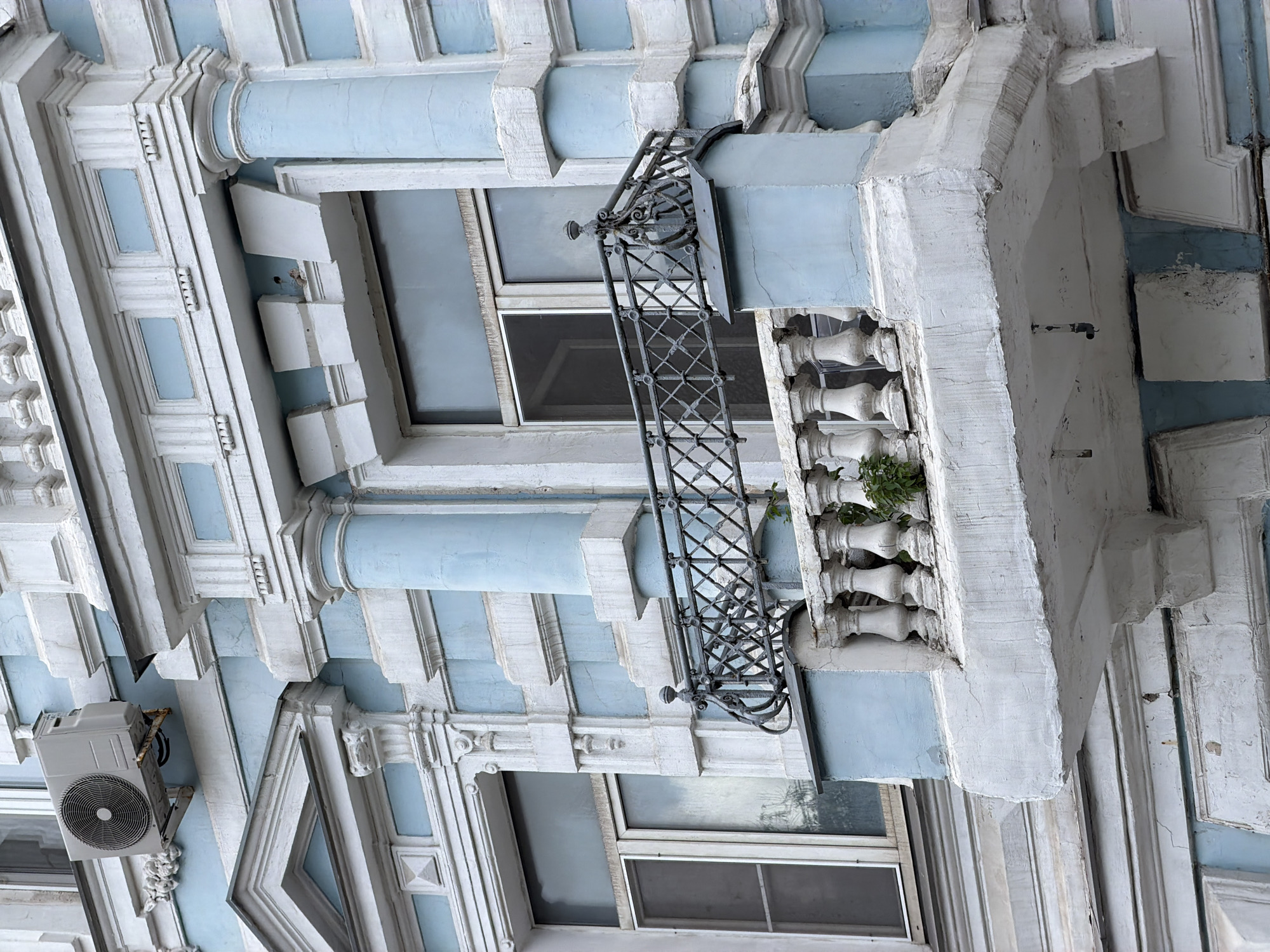 The image shows a close-up, eye-level, exterior shot of a light blue and white building facade with ornate architectural details. A balcony with a decorative metal railing and a balustrade is prominent in the foreground. The balcony appears to be made of concrete and shows signs of age and weathering. There is a small plant growing on the balcony. A window with two panes is visible behind the balcony railing. Above and to the left of the balcony, an outdoor air conditioning unit is mounted on the building wall. The building exhibits classical architectural elements, such as columns, pediments, and decorative moldings. The overall impression is of an older, well-decorated building in an urban setting. This building is likely located in Moscow, Russia, given the architectural style and common use of such building designs in the city. There are no people or animals visible in the image. The environment is overcast or shaded, creating soft lighting.