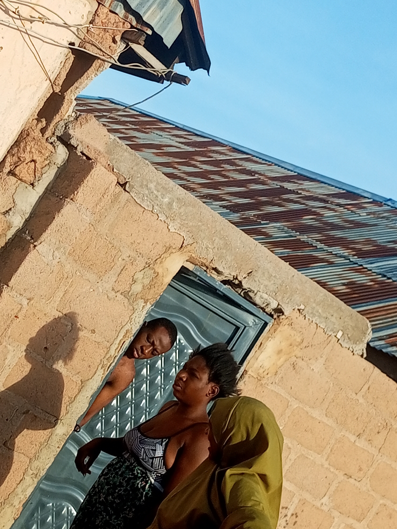 In Tumfure, Nigeria, on a bright, clear day, three individuals are visible in front of a rough, light-brown brick building with a corrugated metal roof. The building's structure appears unfinished or weathered, with exposed wires and chipped plaster. A man is peeking out from an open doorway or window, looking downwards. Beside him, a woman in a patterned dress stands looking to her right, while another person, wearing a green headscarf and garment, is partially visible to her right. The sun casts shadows on the building's textured wall, and the blue sky forms the background.