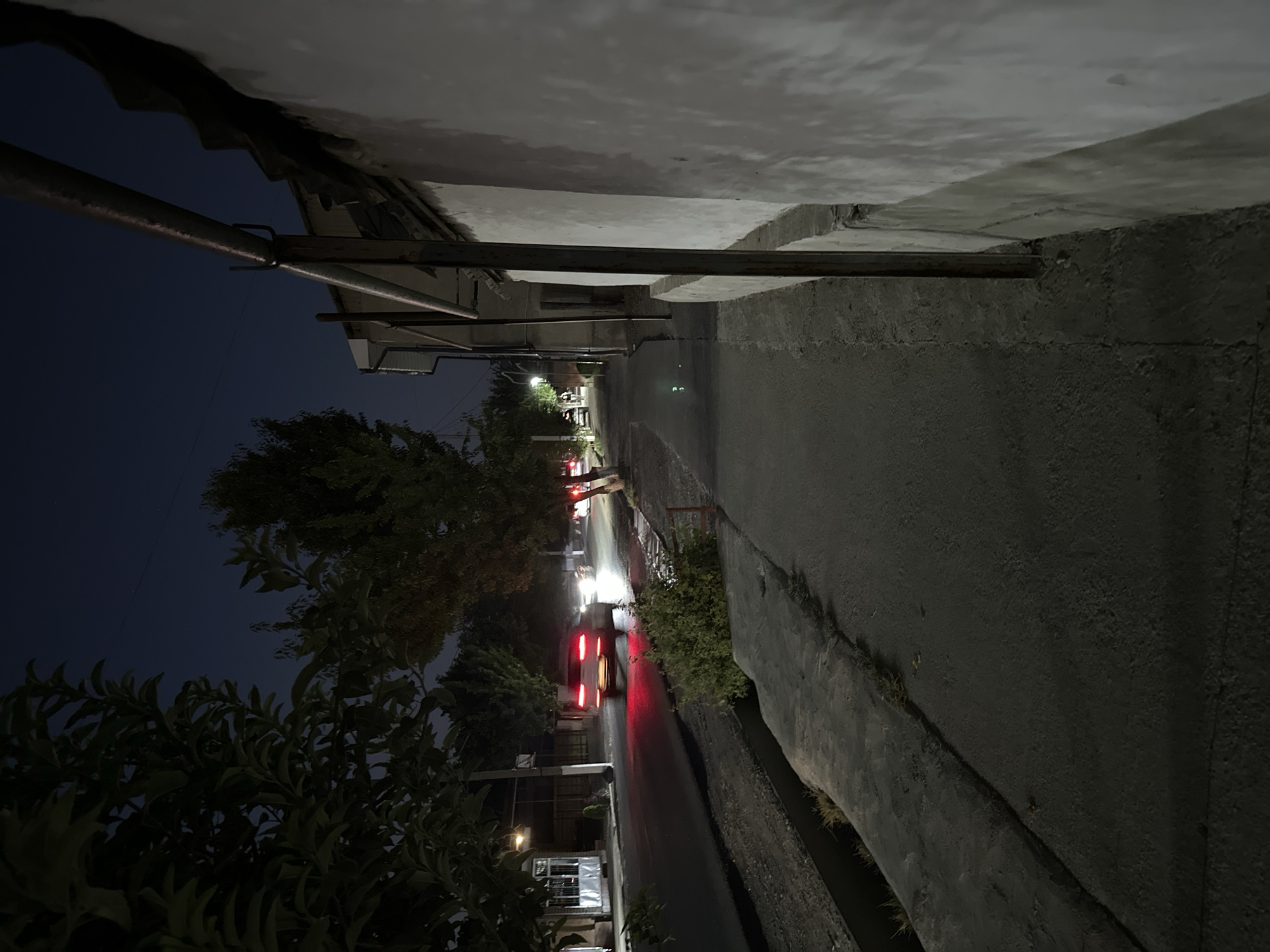 The image depicts a nighttime street scene in Eski-Alimkent, Uzbekistan. A paved sidewalk runs along the right side of the frame, adjacent to a textured, light-colored wall. A metal railing or pipe is visible running parallel to the wall. To the left of the sidewalk is a road with a drainage ditch between the sidewalk and the road. A car with illuminated taillights is driving on the road from left to right, leaving a trail of red light reflections. Trees with dark foliage line the left side of the road. The sky is dark blue, indicating nighttime. The overall impression is of a quiet, residential street at night.