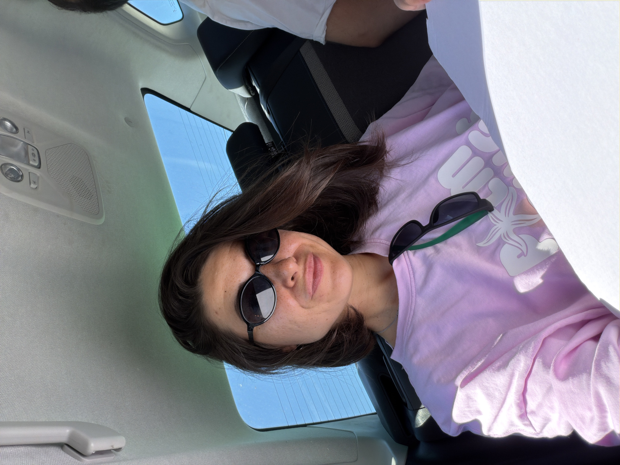 A woman with long dark hair, wearing dark sunglasses and a light pink long-sleeved shirt, is featured in a close-up shot taken inside a car. The image is oriented sideways, with the woman looking towards the camera with a neutral to slightly smiling expression. Her pink shirt has a white tropical-themed graphic, possibly spelling "Hawaii" and including a flower design. A silver chain necklace is visible around her neck.
The setting is the interior of a car, with a light-colored headliner and an overhead console featuring buttons and a speaker grille visible above her. A car seat in a dark material is partially seen in the background. Through the car's rear window, a clear blue sky with faint horizontal lines (likely defroster) is visible, indicating daytime and good weather. Bright sunlight illuminates the scene. A large piece of white paper or fabric is held up near the woman's right side, partially obscuring the frame. The location is Kubasi, Montenegro.