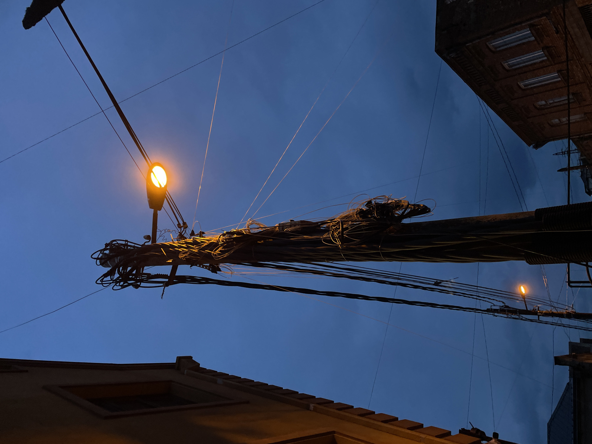 The image captures an urban scene at dusk or night, looking upwards towards a deep blue sky. A prominent utility pole runs diagonally from the bottom-left to the top-right, heavily laden with a thick, tangled mass of black electrical wires and communication cables. A bright, warm yellow-orange street light is affixed to this pole, illuminating the wires immediately around it.
In the distance, further to the right, a smaller, similarly lit street lamp is also visible. Overhead, numerous thinner wires stretch across the sky, some catching the light and appearing as thin, glowing lines.
Parts of two buildings frame the scene: a yellowish-brown roofline with a dark rectangular window is visible in the bottom-left, dimly lit by an unseen source, suggesting a cozy, older structure. On the far right, the upper portion of a darker, taller building with multiple rectangular window frames can be seen, its architectural style hinting at an older urban environment.
The overall impression is one of dense urban infrastructure, characteristic of cities like Tbilisi, Georgia, where such overhead wiring is common. No people or specific activities are depicted, and no visible text is present. The low-angle perspective emphasizes the overhead wires and the evening sky.