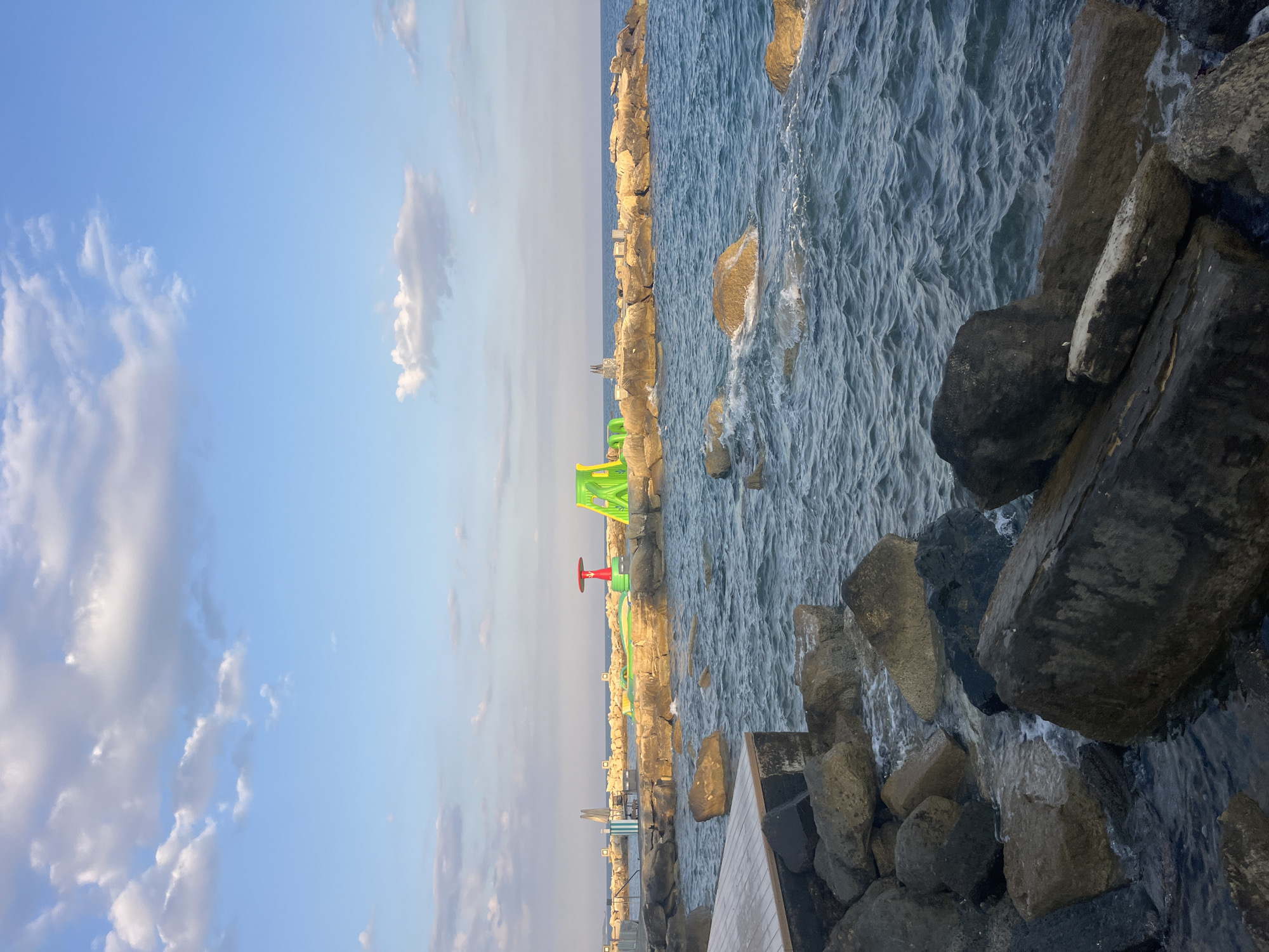 The image depicts a section of the coastline in Agios Tychonas Community, Cyprus. In the foreground, large, dark rocks are submerged in the choppy, dark blue sea. A wooden platform is visible on the left, also with dark rocks around its base. Beyond these rocks, a breakwater made of lighter-colored stones extends into the sea.
On the breakwater, several inflatable water structures are present. To the right, a large, bright green inflatable slide or obstacle course is prominently displayed, with a red and yellow structure next to it. Further to the left on the breakwater, what appear to be lounge chairs or seating areas are visible, along with some smaller green inflatable elements. The sea continues beyond the breakwater, meeting a clear blue sky with scattered white clouds. The scene suggests a recreational area by the sea.