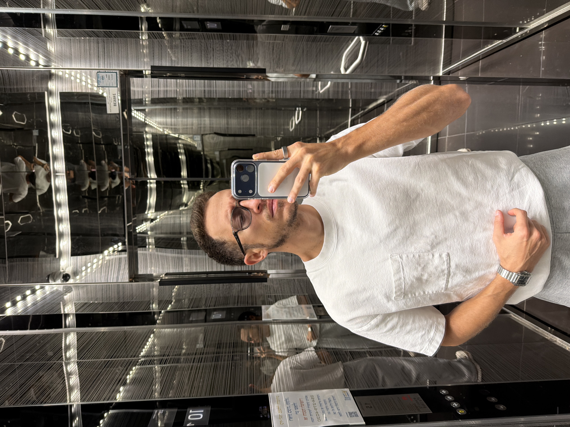 A man is taking a selfie in an elevator. He is wearing glasses, a white t-shirt, and a silver watch. The elevator has mirrored walls and dark metal accents. There are rows of vertical, textured panels that reflect the lights on the ceiling. There are buttons on the elevator panel near the floor. There is also a white information placard attached to the elevator panel. The location is Unknown, Unknown.