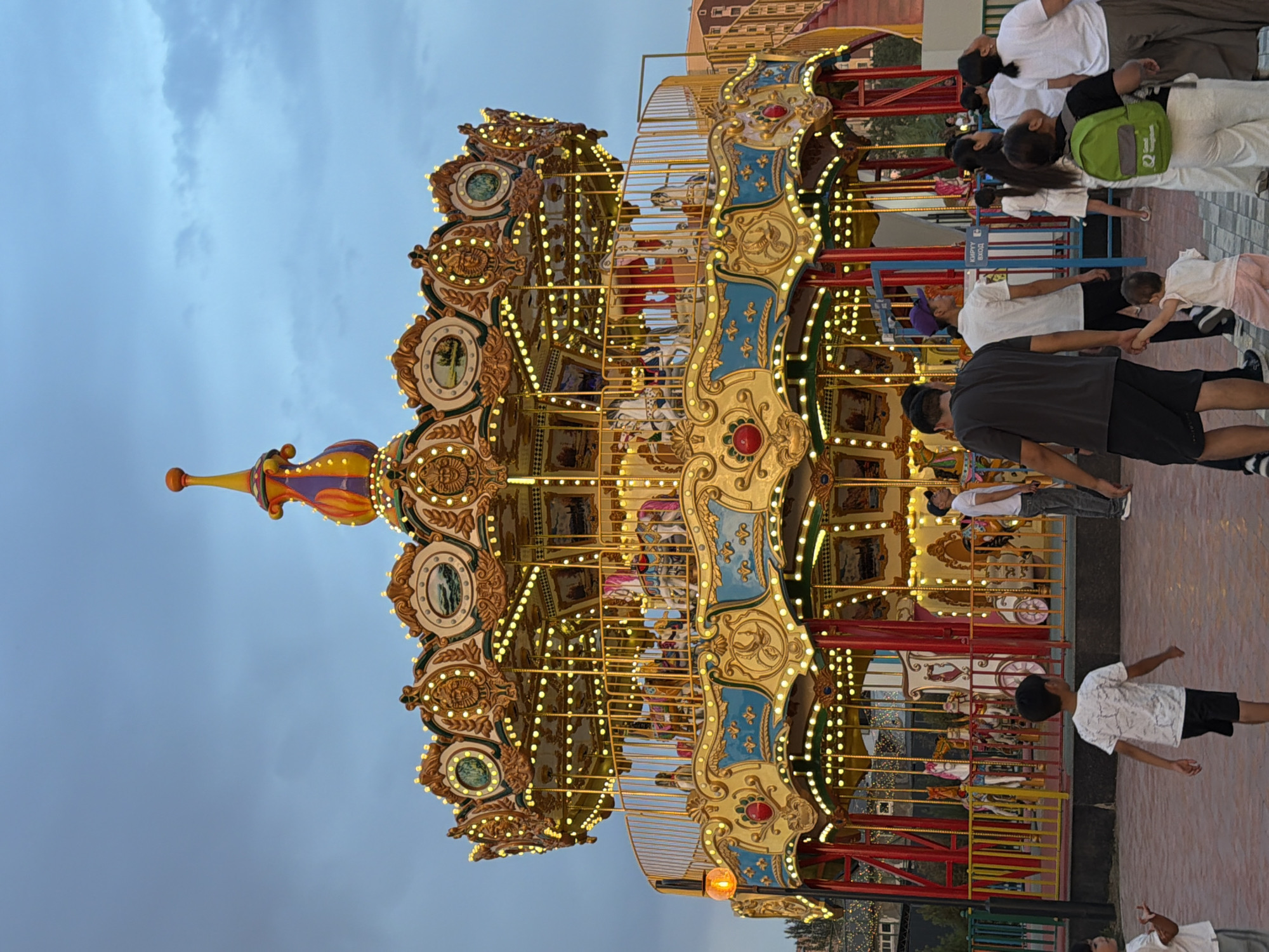 An outdoor amusement park scene at dusk features a brightly lit, ornate carousel as the central focus. The carousel has multiple tiers adorned with gold and blue detailing, a central spire, and numerous small yellow lights illuminating its decorative elements and the painted horses within. Several people, including adults and children, are visible in the foreground and around the carousel. Some individuals are walking, while others appear to be waiting or observing. The ground is paved with brick-like patterns. The sky above is a deep blue, indicating the transition from day to night. In the background, buildings are visible, suggesting an urban or suburban setting. The overall atmosphere is lively and family-oriented.