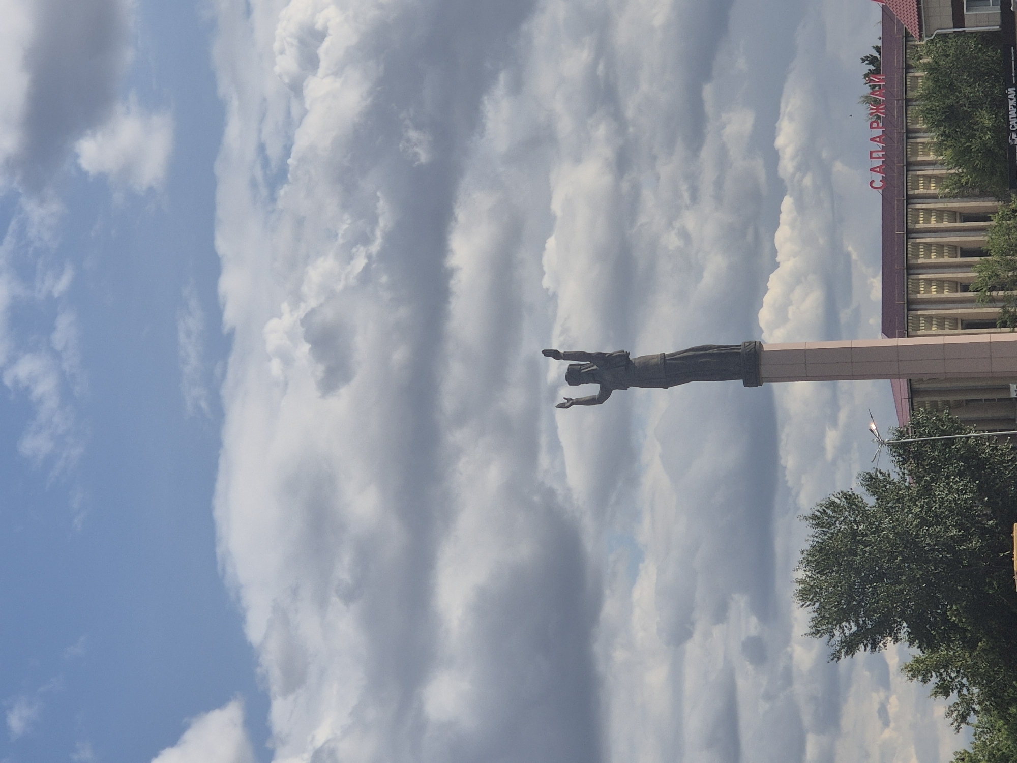 The image captures a statue of a woman with her arms raised, standing atop a light-colored pedestal. Behind the statue, a building with a maroon roof and ornate facade is visible, featuring the Cyrillic text "САПАРЖАЙ" in red letters across its roofline. To the left of the statue, a lush green tree dominates the foreground, with a street lamp visible to its right. The sky is filled with a dramatic mix of blue sky and dense, grey clouds. This scene is located in Kokshetau, Kazakhstan, specifically in front of the Saparzhai building.