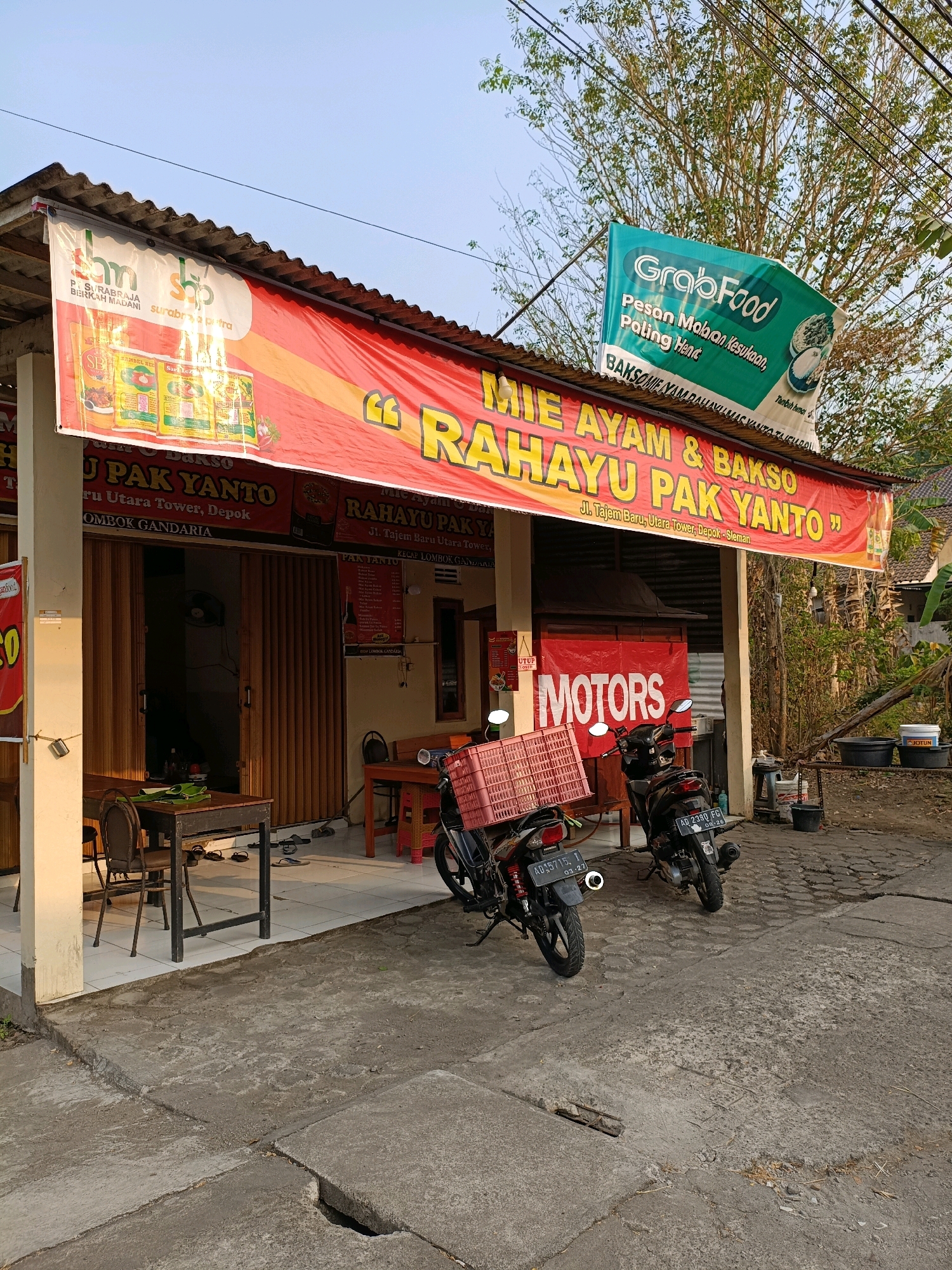 The image shows the exterior of a food establishment in Maguwoharjo, Indonesia. A prominent red banner displays the text "MIE AYAM & BAKSO RAHAYU PAK YANTO" along with the address "Jl. Tajem Baru, Utara Tower, Depok, Sleman". A green GrabFood banner is also visible. Two motorcycles are parked in front of the establishment. One motorcycle has a red crate attached to the rear. Tables and chairs are set up outside, and the entrance is a series of sliding doors. The overall scene depicts a casual dining setting on a clear day.