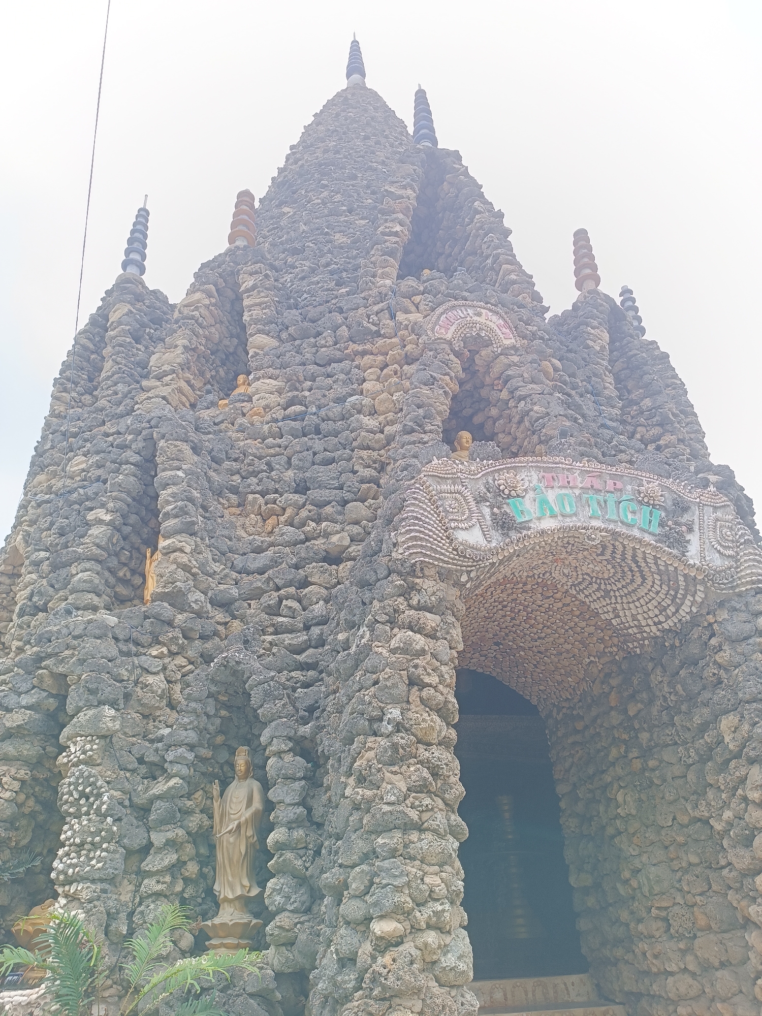 The image shows a large, intricately constructed structure, likely a Buddhist temple or pagoda, located in Cam Ranh, Vietnam. The structure is covered in rocks and seashells, giving it a unique, textured appearance. At the peak of the structure are several decorative spires. The entrance to the structure is an arched doorway adorned with carvings and a sign with text in Vietnamese, appearing to read "THAP BAOTICH". Inside the entrance, a dark interior with some golden statues is visible. On the left side of the structure, a statue of a standing figure, possibly a deity, is situated within a rocky alcove, with some greenery at its base. The overall impression is one of a visually striking and culturally significant religious site.
