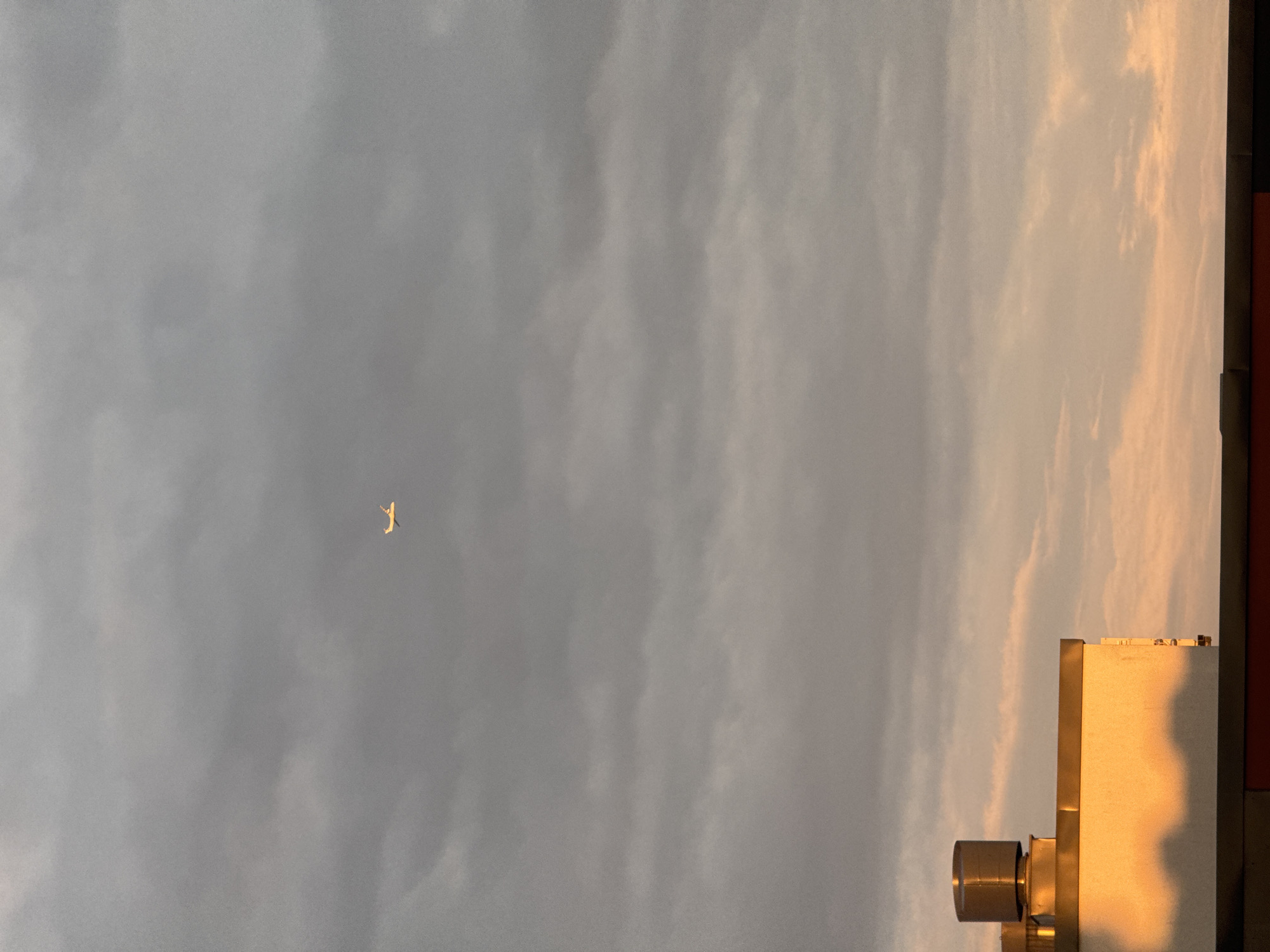 This media file shows a view of a cloudy sky with an airplane in the distance. The left side of the image is dominated by gray clouds. On the right side, the clouds are tinged with a warm, orange-pink hue, suggesting it is either sunrise or sunset. Partially visible in the bottom right corner is the side of a building, catching the warm light. The building has a metallic cylindrical feature and appears to be modern. The airplane is small and silhouetted against the sky, appearing to be in flight. The overall mood is serene, with the soft light creating a peaceful atmosphere. There is no visible text in the image.