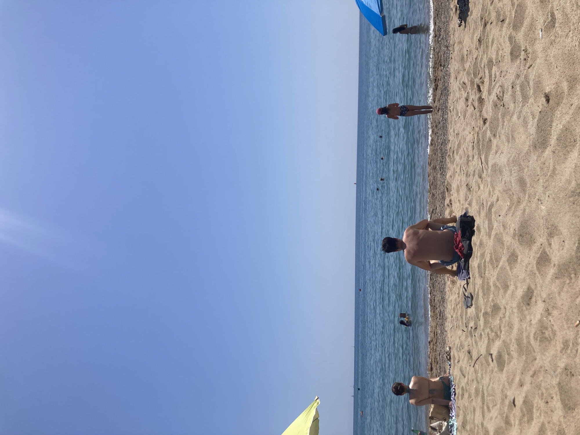 The image depicts a sunny day at a beach in Barcelona, Spain. The scene is divided horizontally, with the clear blue sky occupying the left side and the sandy beach and ocean on the right. Several people are visible, enjoying the water and the shore.
In the foreground, a man is sitting on the sand, facing the ocean. He is shirtless and appears to be resting. Further down the beach, another person is seated, also facing the water, wearing a bikini. In the ocean, a few individuals are swimming or wading in the water. One person, wearing a pink hat, is standing in the shallow water. Farther out, more people are scattered across the sea.
Objects visible include a blue beach umbrella, a yellow umbrella in the lower left corner, and some bags and towels on the sand. The sand is light brown and textured, with small pebbles near the waterline. The ocean is a clear blue, with gentle waves lapping the shore.
The time of day appears to be daytime, likely late morning or early afternoon, given the bright sunlight and clear sky. The weather seems warm and pleasant, typical of a beach day. The presence of people in swimwear and the general atmosphere suggest a leisurely recreational activity. No text is visible in the image.