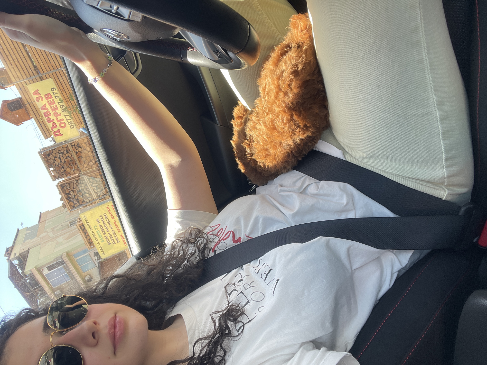 The image shows a woman wearing sunglasses and a white t-shirt with a seatbelt, sitting in the driver's seat of a car. She is holding the steering wheel. On the passenger seat, there's a small, furry, brown dog next to a white fabric-covered item or object. A bracelet adorns the woman's wrist. Outside the car window, a building with Bulgarian text on a sign is visible. The text includes "СКЛАД ЗА ДЪРВЕН МАТЕРИАЛ БАШЕВ-73" (Warehouse for Wood Material Bashev-73) and a phone number "0877 897.779" which is identifiable as a Bulgarian phone number. Another sign reads "ДЪРВА ЗА ОГРЕЕВ". Based on the Bulgarian text, the location is likely in Bulgaria, possibly Sofia.