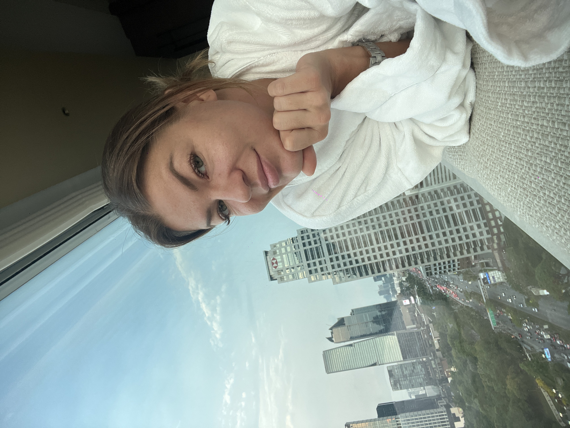 A woman with light brown hair is wearing a white bathrobe and leaning against a light gray upholstered headboard or wall. She is looking directly at the camera with a slight smile. She is wearing a silver watch on her left wrist.
Behind her, a window shows a view of Bangkok, Thailand. There are several high-rise buildings, including one with a red and white logo (likely HSBC). Below the buildings, a highway is visible with moderate traffic. The sky is blue with some clouds.
