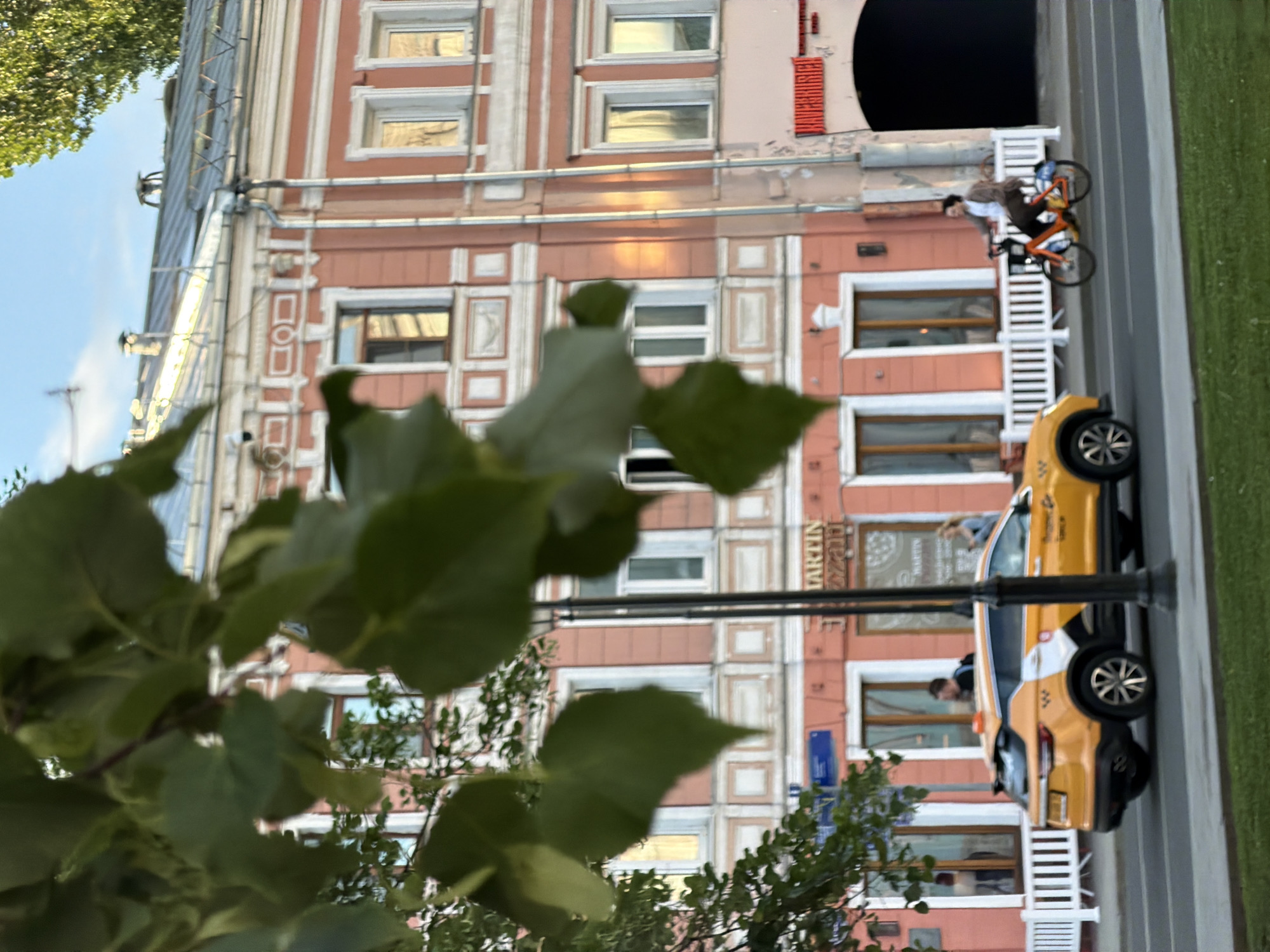 The image depicts a street scene in Moscow, Russia, focusing on a section of a pink building with white architectural details and window frames. A yellow taxi is parked on the street, and a person is riding an orange bicycle on the sidewalk. Lush green foliage is visible in the foreground, partially obscuring the view. The architecture suggests an older part of the city, potentially a historical district. The presence of a taxi and a cyclist suggests a modern urban environment.