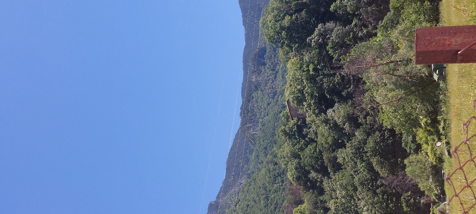 The image shows a landscape in Bachkovo, Bulgaria. A mountain covered with dense green trees rises on the right side of the frame. A small structure is nestled among the trees on the mountainside. Overhead power lines run along the slope of the mountain. The left side of the image features a clear blue sky. In the foreground, a grassy area and a metal fence are partially visible, obscuring the lower right corner of the scene. A brown, rusty metal beam or structure is present in the right foreground.