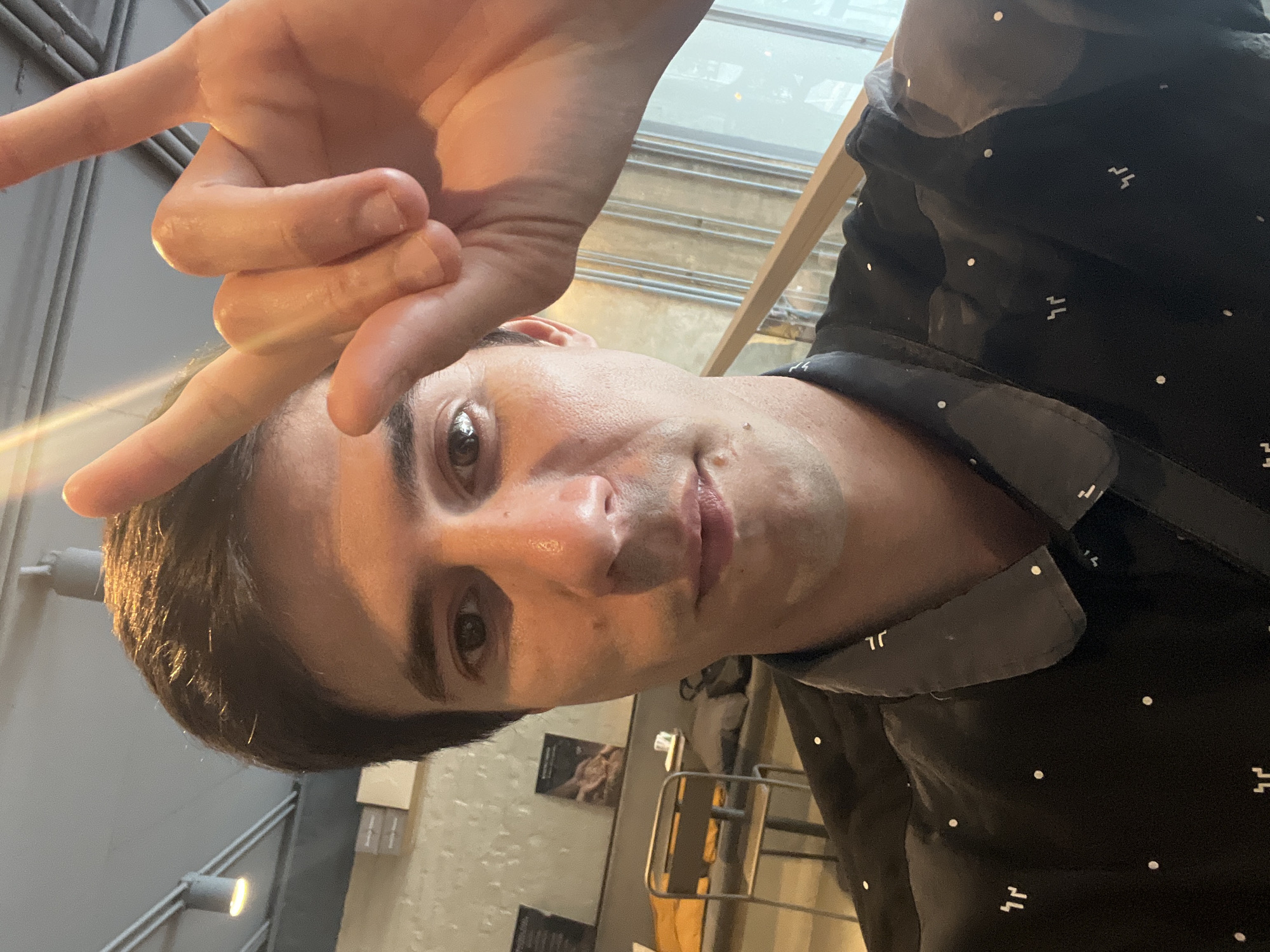 A selfie of a man with dark hair, light skin, and brown eyes, facing the camera. He is wearing a black patterned shirt with a gray collar. His right hand is raised above his head, making a hand gesture. The background is blurred but shows a wall with posters or pictures, and metal structures. The setting appears to be indoors, possibly a restaurant or bar. The location cannot be determined to be Buenos Aires without additional information.