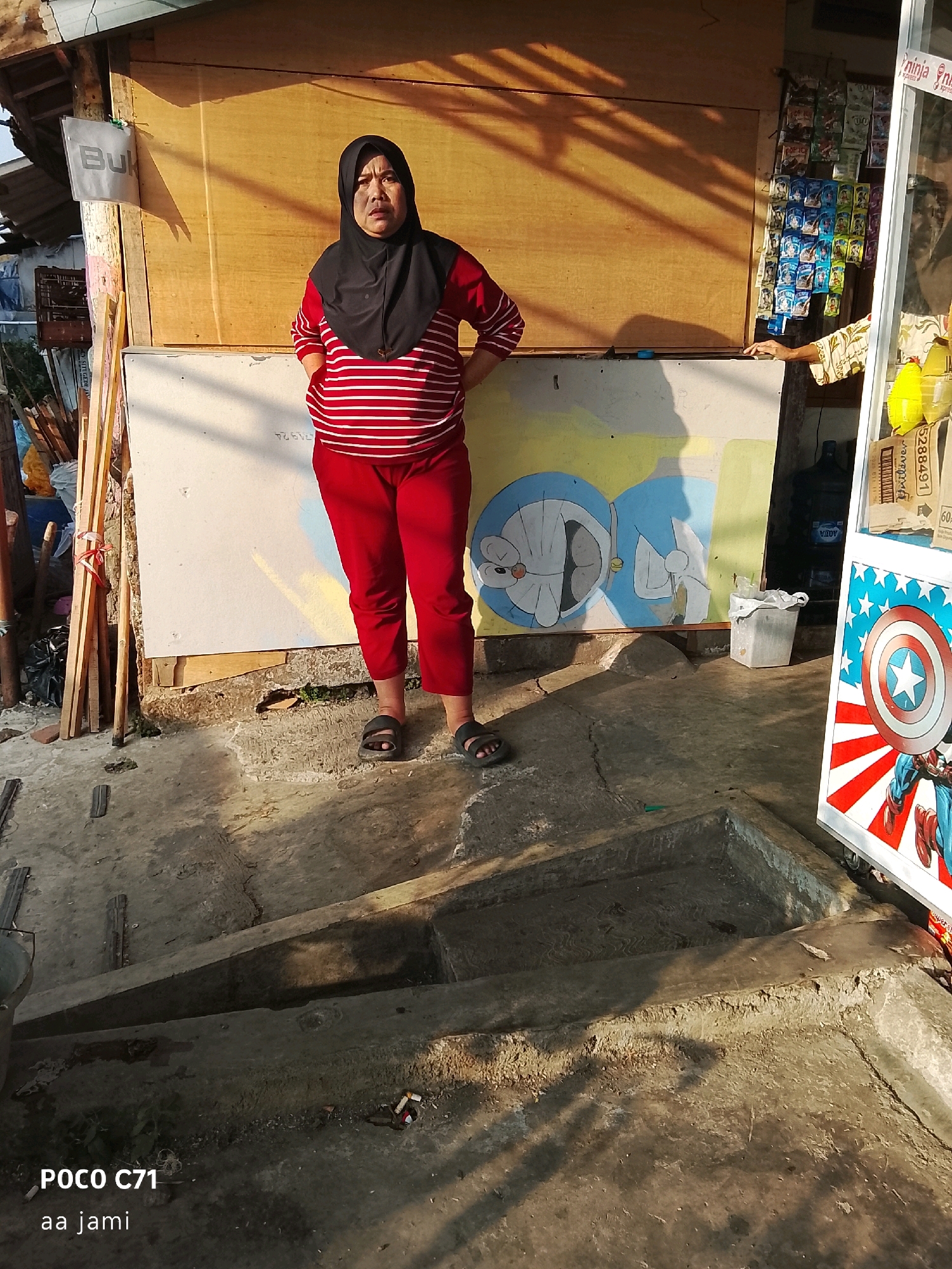 A woman wearing a black hijab and a red and white striped long-sleeved shirt stands facing forward, her hands on her hips, in front of a wooden wall and a large white board with a painting of Doraemon on it. She is wearing red pants and black sandals. To the right of the woman, a display rack holds various small packets of products, and a part of a white display board with a Captain America shield graphic is visible. In the background, stacks of wood and debris are present. The ground is concrete and appears uneven, with a small set of steps. The scene is outdoors, illuminated by sunlight casting shadows. The image is identified with the text "POCO C71 aa jami" at the bottom left. The location is identified as a street stall in Tasikmalaya, Indonesia.