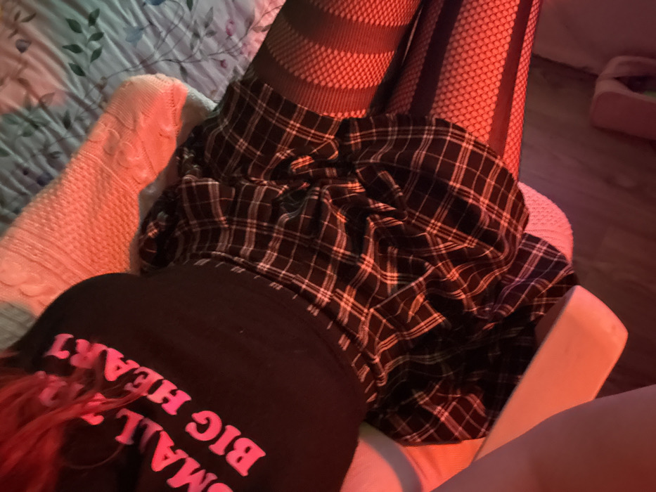 The image presents a close-up, downward-angled view of a person's upper torso and legs, positioned on a padded surface, likely a bed. The individual's hair, appearing reddish due to ambient lighting, is visible at the top. They are wearing a black sleeveless top featuring the bright pink capitalized text "SMALL T BIG HEART." Below the top, a dark plaid skirt with black and thin white intersecting lines and a ruffled or pleated texture is visible. Both legs are covered in dark hosiery; the right leg shows a consistent fishnet pattern, while the left leg displays alternating horizontal opaque and patterned bands leading into a fishnet design on the lower portion of the thigh.
The immediate environment includes a bedspread with a light background and a subtle floral or leafy pattern in muted blue and green tones. A cream or white colored cable-knit textured item, possibly a pillow or blanket, is situated beneath the person's upper body. The scene is illuminated by prominent red ambient lighting, casting a strong reddish-pink hue across the subject, the cable-knit item, and parts of the bedspread, particularly on the right side of the frame. A faint greenish or bluish tint is also present in some areas. A portion of a white, rigid object, possibly a piece of furniture, is visible in the lower left corner. The specific location within the city of Yerevan, Armenia, cannot be determined from the visual information presented in the image.