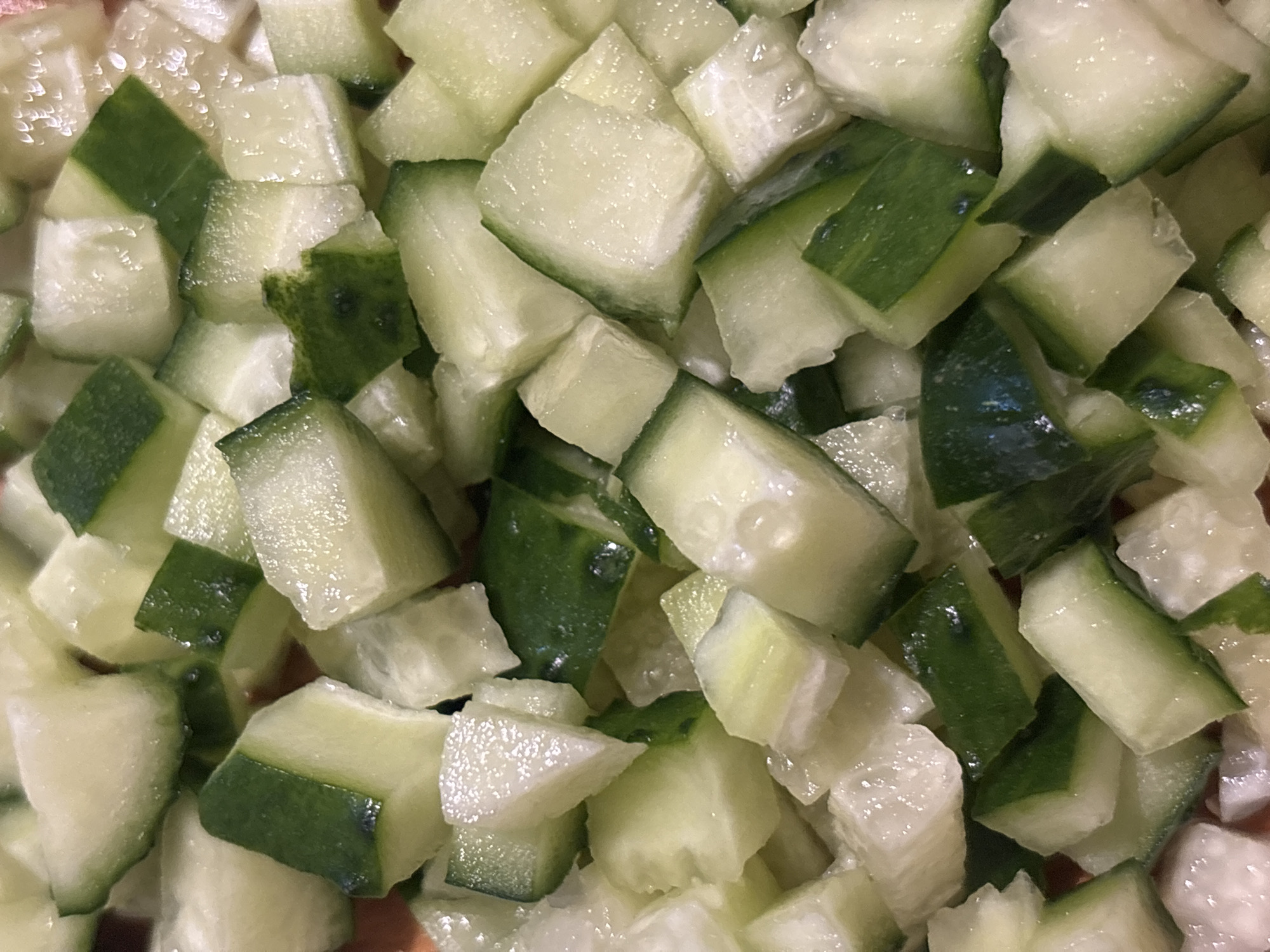 A close-up, overhead shot shows a pile of chopped cucumbers. The cucumbers are diced into irregular, bite-sized pieces, with visible green skin and white flesh. Some pieces are wet, creating small glistening highlights. The overall color palette is predominantly green and white, with hints of the orange surface beneath the cucumbers peeking through in some areas. The image provides no context related to the city of Kremenchuk, Ukraine, nor does it depict any people, actions, or interactions. It is solely a visual representation of diced cucumbers.