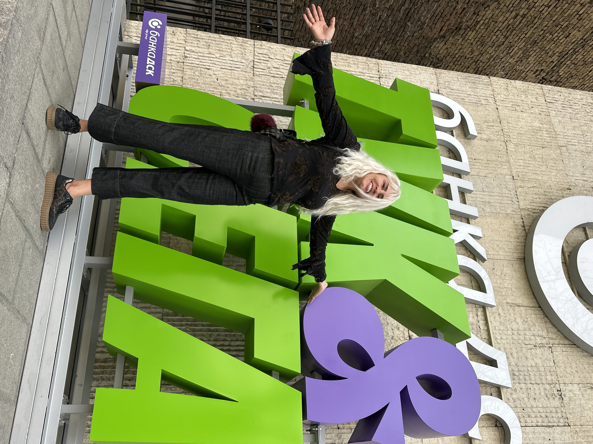 A woman poses in front of the Bank DSK sign in Sofia, Bulgaria. She has long white hair, wears a black top with a floral design, dark jeans, and black platform shoes. She stands on the sign with one hand raised, smiling. The sign is constructed of large, green, purple, and white letters. The background includes a concrete wall and a metal grate.