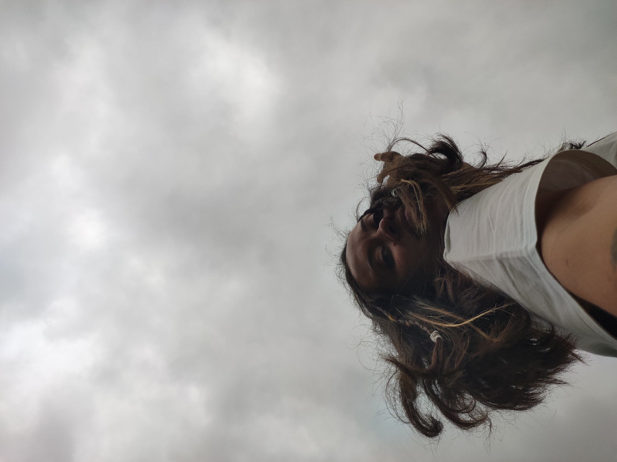 A low-angle upward-facing photographic image, captured in Фоки, Russia, depicts the head and upper torso of a human individual against an overcast sky. The subject is positioned in the lower right portion of the frame, viewed from a worm's-eye perspective, with their head inverted relative to a standard upright orientation. The individual has long, dark, wavy hair, with some strands appearing wild or frizzy. Small braids or twisted sections of hair with light-colored beads or wraps are visible. The subject wears a light-colored (likely white) short-sleeved garment on their right shoulder and arm. Portions of the face are visible, including the nose, mouth area, and one eye, partially obscured by hair and shadows due to the upward angle of the shot. A dark marking or tattoo is faintly discernible on the upper right arm/shoulder area. The background consists entirely of a cloudy sky, presenting a palette of grays and whites, indicating an overcast atmospheric condition. No ground features or structures are visible.