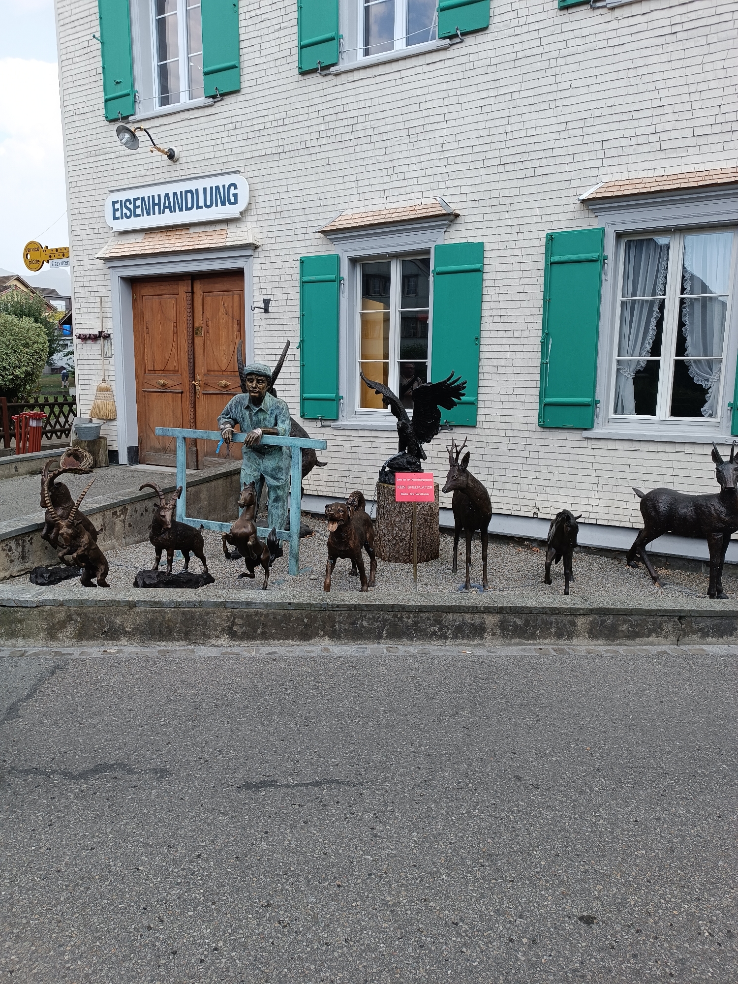 In the city of Appenzell, Switzerland, a storefront displays the sign "EISENHANDLUNG," indicating a hardware or ironmongery business. The building's facade is constructed of light-colored brickwork, adorned with vibrant green shutters on its windows. In front of the entrance, a collection of bronze animal sculptures and a human figurine are arranged along a low wall bordering the street. The human figure, depicted as a man in overalls and a cap, leans against a blue railing, holding a tool, possibly a brush, in his hand. Surrounding him are various animal sculptures, including what appear to be ibexes, a deer, a dog, and a bird of prey perched on a stand. A red sign is placed on the bird of prey's stand. A paved sidewalk runs in front of the sculptures, leading to the street, which is made of asphalt. The scene suggests a display of artwork or merchandise outside the business, set against the backdrop of a Swiss town.