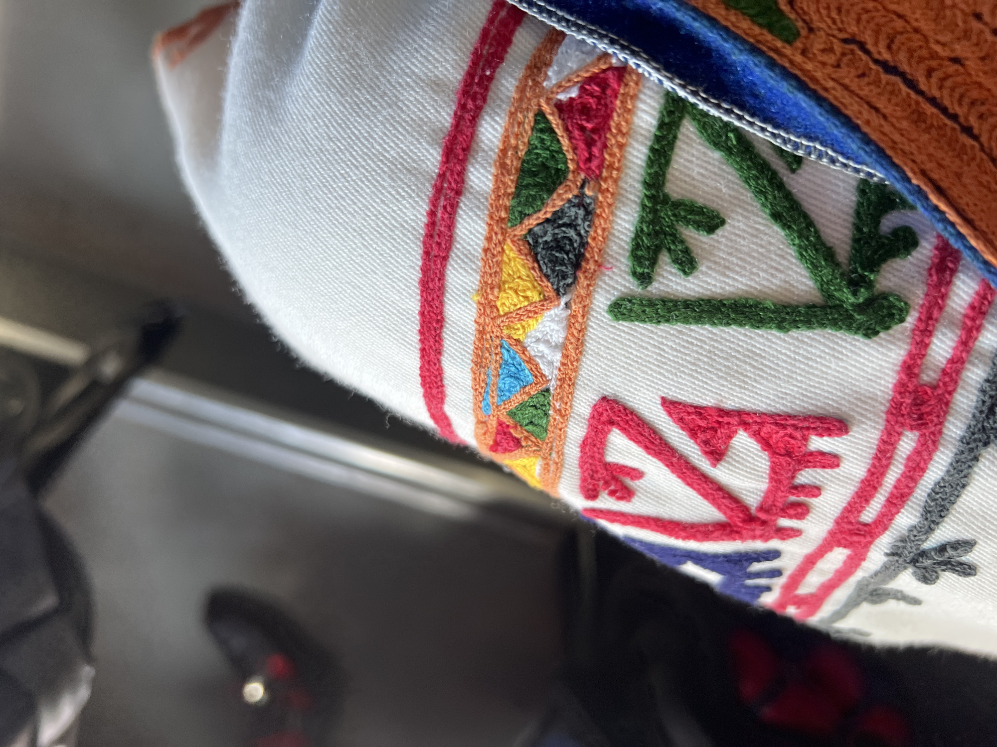 The image displays a close-up, angled perspective of a white textile adorned with elaborate, multi-colored embroidery, situated in an interior setting in Slivovo, Bulgaria. The primary subject is a section of off-white fabric, exhibiting a visible diagonal weave.
Three horizontal bands of embroidery are prominent. The top band consists of a narrow, solid line of bright red stitching. Below this, a wider band features an orange embroidered outline enclosing a series of contiguous, filled-in triangles. These triangles present a mosaic of colors, including blue, yellow, dark green, white, black, and red. Further down on the white fabric, larger, abstract geometric patterns are embroidered in dark red, royal blue, dark grey, and dark green threads. These patterns are characterized by angular lines with short, perpendicular extensions.
On the far right, a vertical strip of blue fabric trim is visible, partially obscuring an adjacent textured, orange-colored material. The background is out of focus and dark, suggesting ambient or low light conditions indoors. Vague shapes and light reflections are discernible in the blurred background, including a dark, amorphous form in the upper left and a blurred reddish object with a bright, circular reflection in the mid-left. No individuals are depicted in the scene.