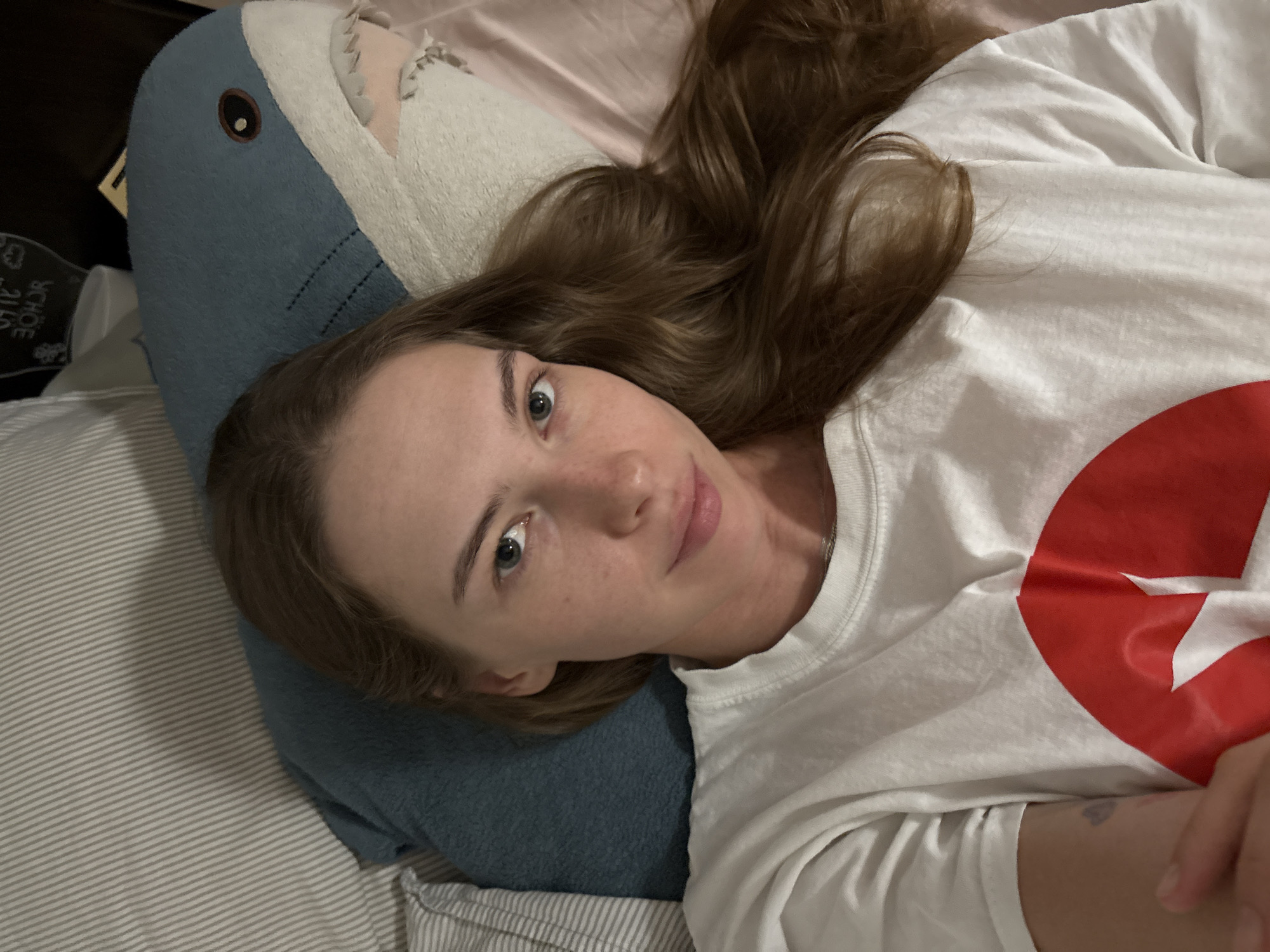 A young Caucasian woman with long, brown hair is lying on a bed, partially obscured by a large blue shark plush toy. She is wearing a white t-shirt with a large red graphic on the front. The bed appears to be in a bedroom, with striped pillows and a pink sheet visible. The scene is dimly lit, suggesting it may be nighttime or indoors with low lighting. The woman is looking towards the camera with a neutral expression. There is no information available to determine the location in the city of Perm, Russia.