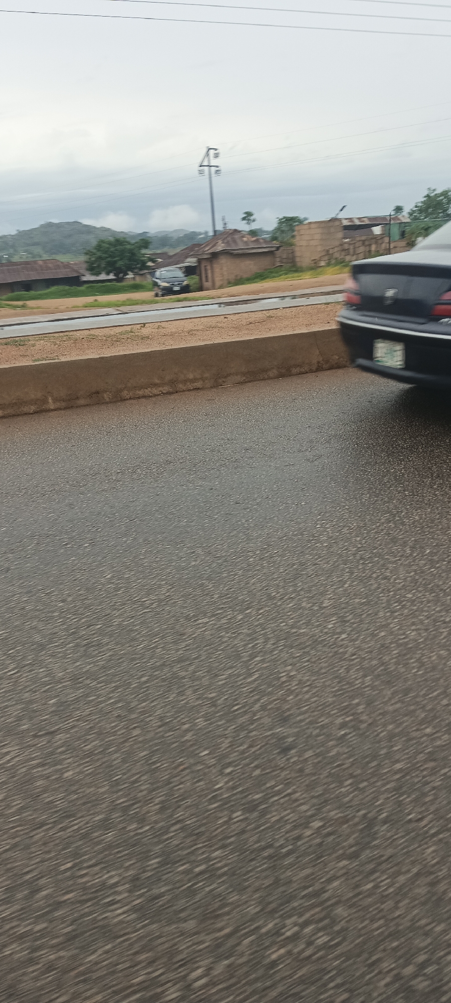 The image captures a roadside scene in Bukuru, Nigeria, under an overcast sky, suggesting either a cloudy day or recent rainfall, as the asphalt road in the foreground appears wet and somewhat reflective. The perspective is from a moving vehicle, indicated by the slight motion blur on the road surface.
In the immediate foreground on the right, the rear of a dark-colored sedan is partially visible, featuring a license plate that is indistinct due to blur and distance. A concrete curb or barrier runs horizontally across the middle ground, separating the main road from an elevated unpaved shoulder.
Beyond this barrier, scattered single-story buildings are visible. These structures appear to be constructed from materials like mud bricks or concrete blocks and are topped with corrugated iron roofs, typical of local architecture in some parts of Nigeria. Among these buildings and sparse vegetation, a dark SUV or minivan can be seen parked or moving slowly on the unpaved area on the left side of the frame. A tall metal utility pole with cross-arms and a street light fixture stands towards the right of the background, with power lines extending across the upper portion of the image. Hilly terrain, partially obscured by the hazy, cloudy conditions, forms the distant background. No people are visible in the frame. The scene conveys a quiet, semi-urban or rural environment.