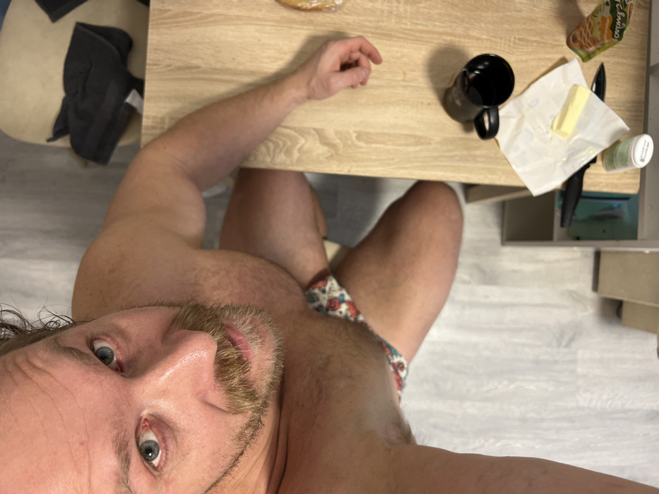 A shirtless man takes a selfie from a low angle while sitting at a table. The table holds a cup, butter, a knife, and other items. He wears patterned underwear. Clothes sit on a chair nearby. The floor is visible.