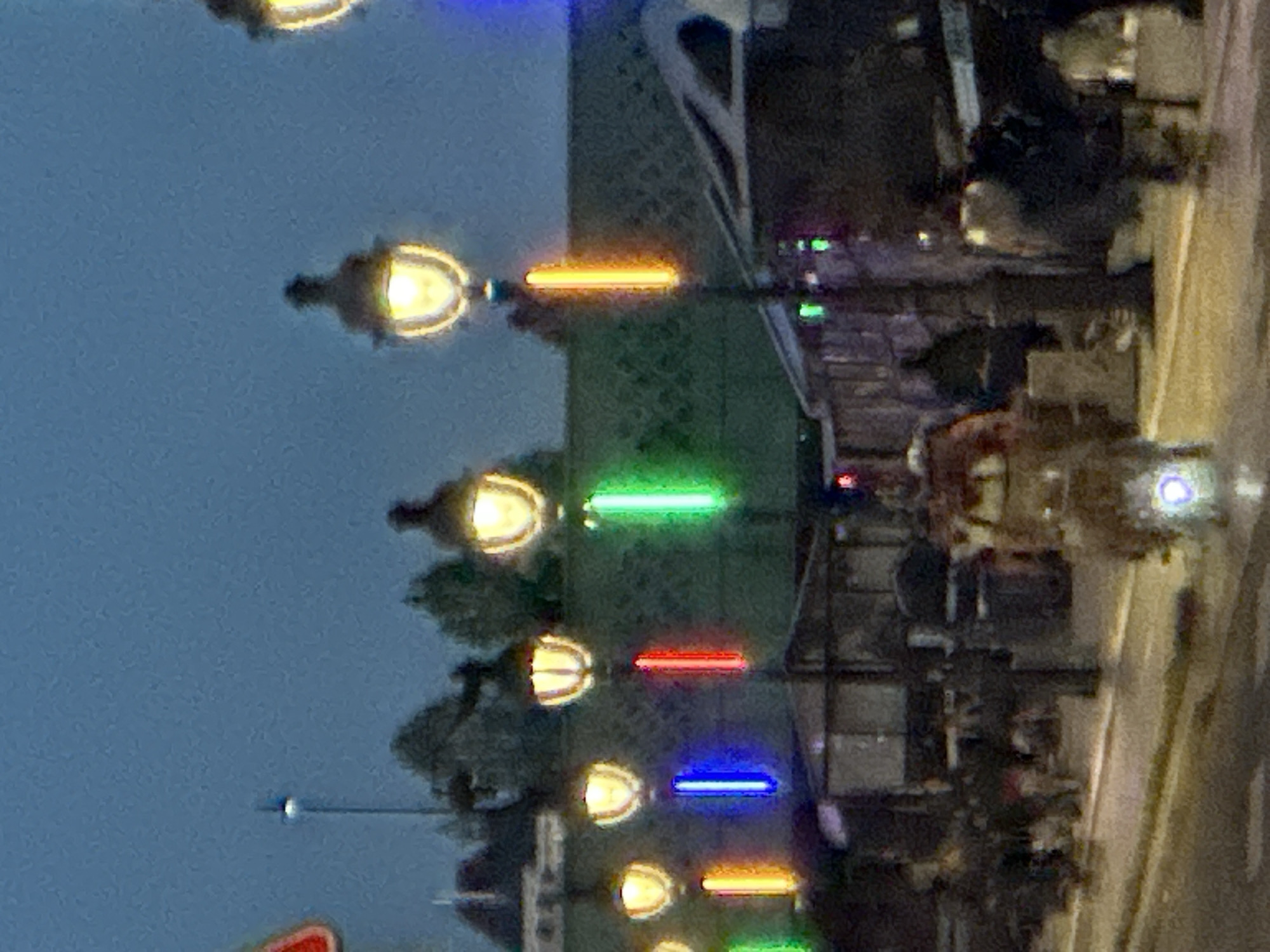 The image depicts a street scene in Paris, France, at dusk or night. The scene is dominated by a row of old-fashioned lampposts illuminated with various colors (orange, green, red, blue). These are likely located along a prominent boulevard or street. The lampposts are interspersed with trees, suggesting they are positioned along a park or walkway. The background suggests the presence of buildings, potentially restaurants or shops, with tables and chairs set up along the street. The presence of people is implied. The composition suggests a view from above street level. The street is also lit by other lights.