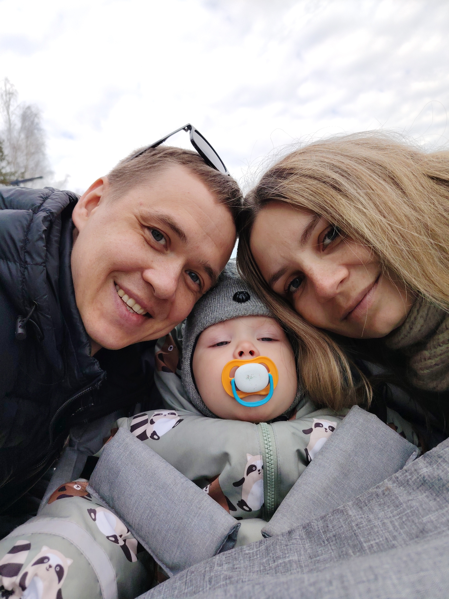 A selfie taken in Chaikovsky, Russia, depicts a family group of three people. A man and a woman are close together at the top of the frame, smiling at the camera. The man has short hair and is wearing a black jacket with sunglasses on his head. The woman has long blonde hair and is wearing a light-colored scarf. Below them is a baby wearing a grey hat and a green jacket with a pattern of raccoons. The baby is resting in a grey stroller or carrier and has a pacifier in its mouth. The background shows a cloudy sky and some trees.