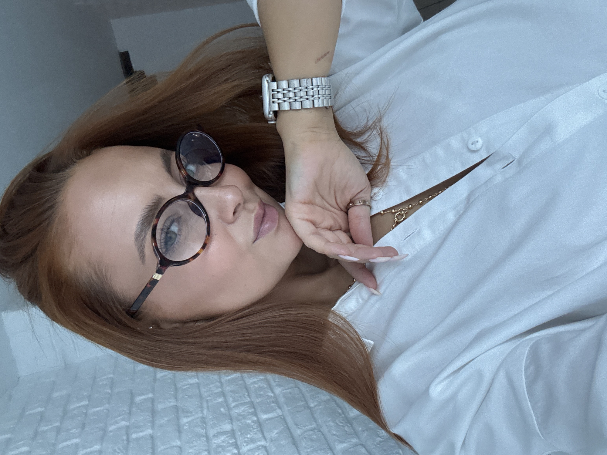 An upward-facing portrait captures a woman with long, reddish-brown hair, styled to her left side. She wears round, tortoiseshell-framed glasses, through which her light-colored eyes are visible. Her lips are slightly parted, and her expression is pensive or contemplative.
She is dressed in a crisp white button-down shirt, unbuttoned at the top to reveal a delicate gold-toned necklace with small beads and a circular charm. On her left wrist, she wears a silver-toned watch with a metal link bracelet, resembling a smartwatch. Her left hand is raised, with manicured fingers lightly touching her jawline and cheek. A gold-toned ring, possibly inscribed with Roman numerals, is visible on her right ring finger.
The background consists of a plain, light-colored surface behind her head, and a textured white surface, possibly a quilted headboard or brick-patterned wall, beneath and to her left. The soft, even lighting suggests an indoor setting. The image portrays a calm and stylish scene. The subject's location is Chaikovsky, Russia.