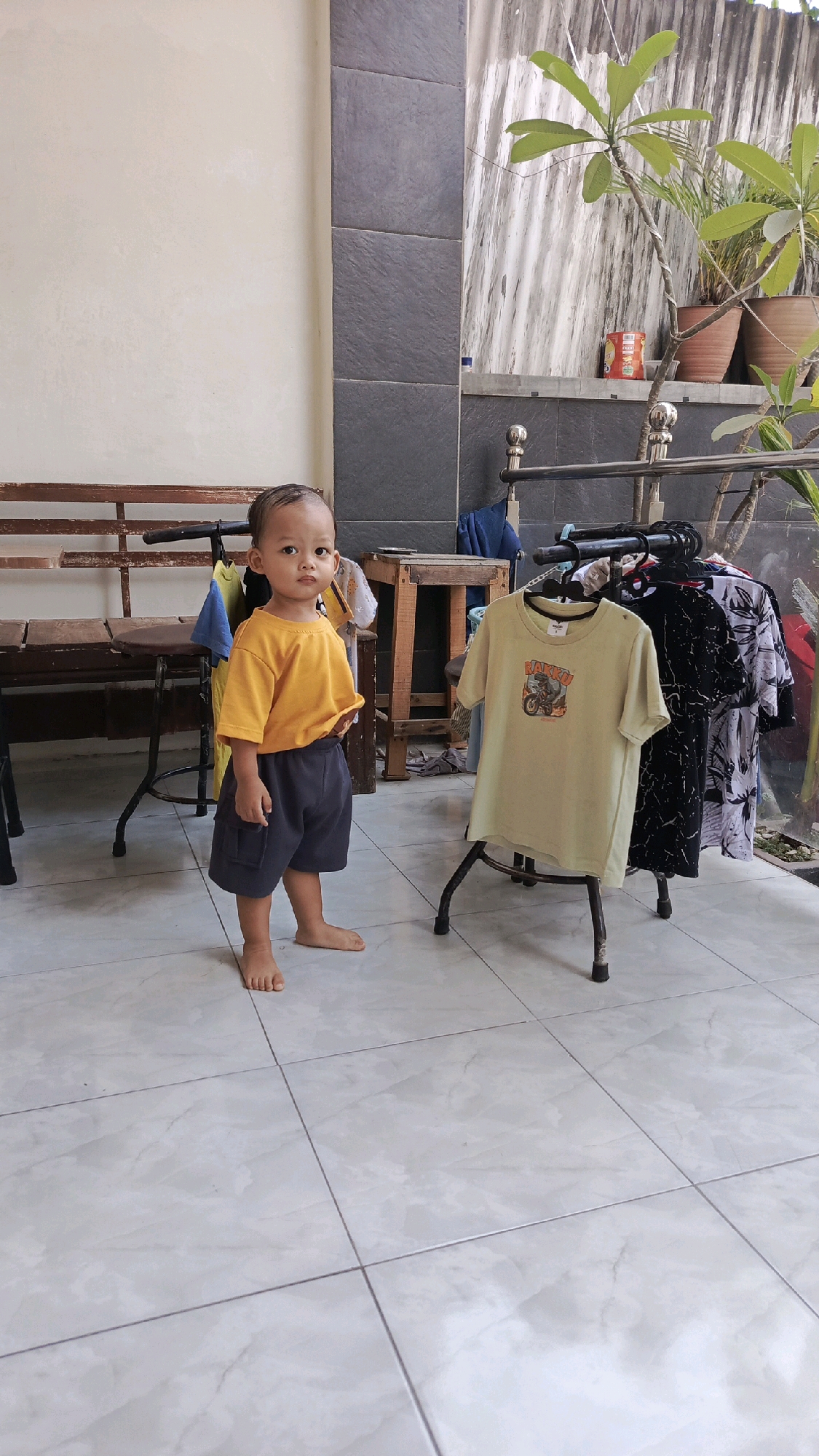 A young Indonesian child stands barefoot on a tiled patio, wearing a yellow t-shirt and dark shorts. The child is positioned in front of a clothes rack displaying several t-shirts, with a wooden bench and a table to their left. In the background, potted plants are visible against a textured wall. The scene appears to be an outdoor area of a residential property.