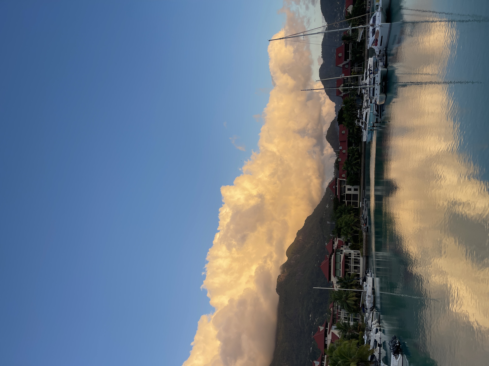 The image depicts a serene marina scene in Victoria, Seychelles, bathed in the warm glow of what appears to be sunrise or sunset. The sky above is a vibrant, clear blue, with a dramatic bank of large, fluffy clouds stretching across the mid-section of the frame. These clouds are tinged with a golden-orange hue, suggesting the low angle of the sun.
Below the clouds, a range of lush, green mountains forms the backdrop. The slopes are partially obscured by mist or low-lying clouds in places, adding to the atmospheric quality of the scene. In the foreground, a tranquil body of turquoise water reflects the sky, clouds, and the surrounding structures. The reflections are clear, mirroring the vibrant colors of the sky and the golden light on the clouds.
Along the waterfront, a collection of buildings with distinctive red-tiled roofs are visible, interspersed with palm trees. These structures appear to be part of a resort or residential complex. A marina is situated in the water, with numerous boats and yachts docked. The vessels are of various sizes, with several sailboats featuring prominent masts. Some smaller motorboats are also present.
The overall impression is one of peacefulness and natural beauty, highlighting the scenic harbor of Victoria. There are no people visible in the image.