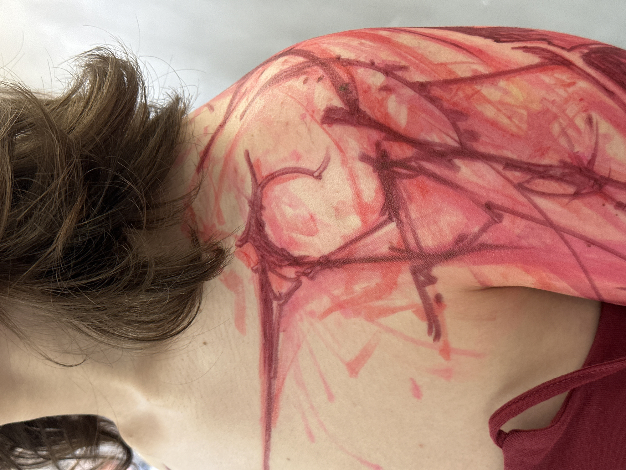 The image shows a person with brown hair lying down. Their upper left arm and shoulder are visible, covered in red and burgundy markings resembling scratches or drawings. The person is wearing a red tank top. The background is a blurry white. No information indicates a location in Moscow, Russia.