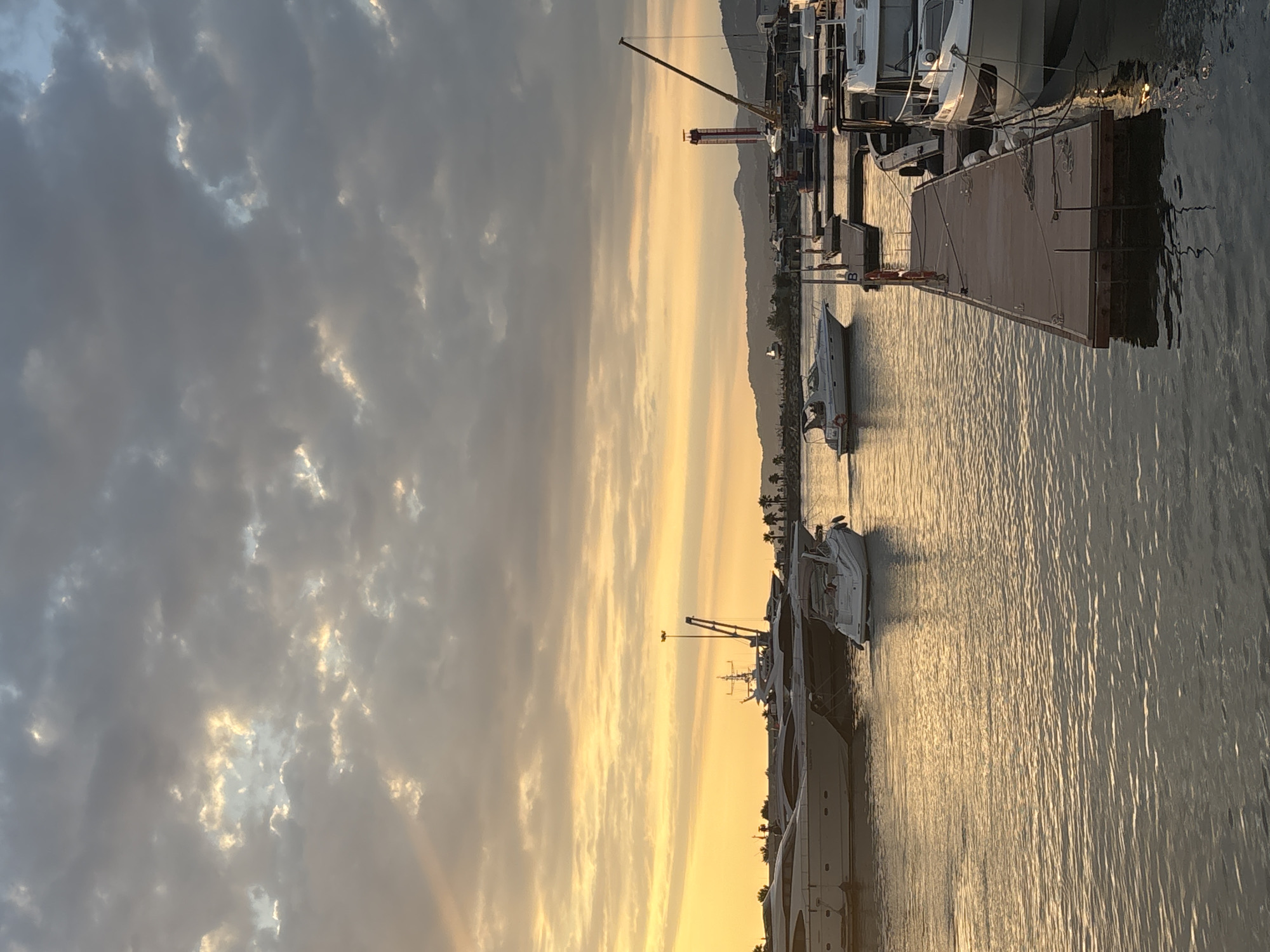 The image depicts a marina in Sochi, Russia, at sunset. The sky is filled with dramatic clouds, with streaks of sunlight breaking through, casting a warm, golden glow on the scene. Several yachts and smaller boats are docked or moving in the water. In the foreground, a wooden pier extends into the water, with a large white boat moored to its right. Further back, more yachts are visible, and a crane stands tall near the shore. In the distance, a line of palm trees and buildings suggests a coastal city. The overall atmosphere is serene and picturesque, capturing the beauty of a Mediterranean sunset over a bustling harbor.