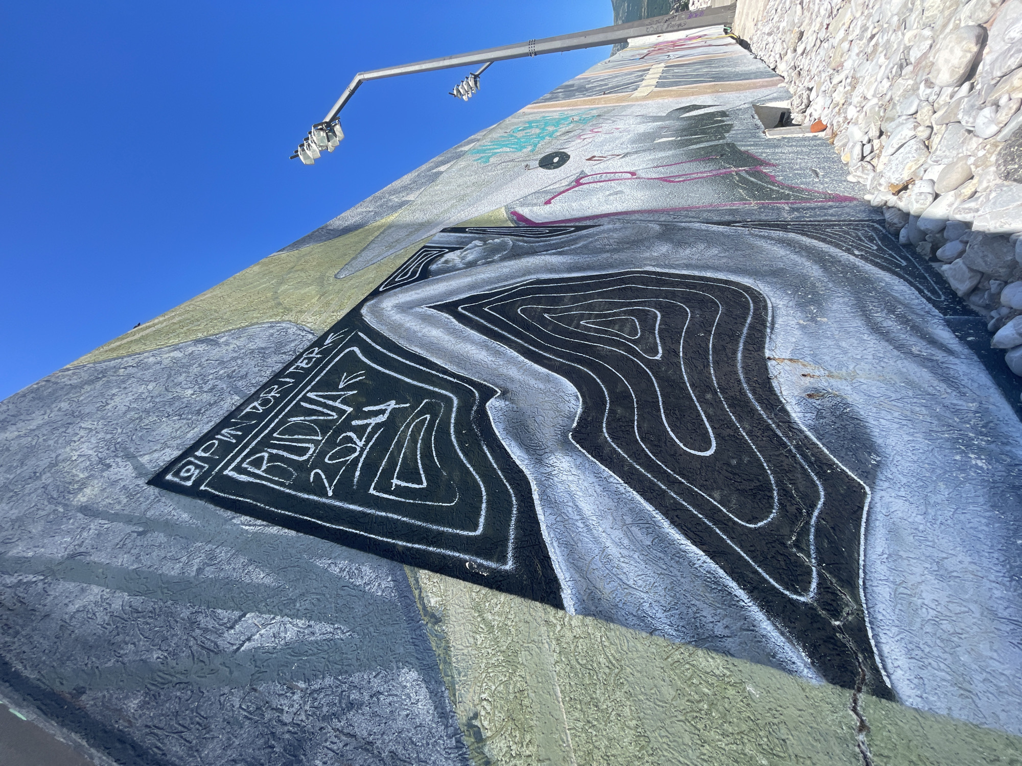 A low-angle, outdoor shot presents a mural on a concrete surface. A light fixture extends into the upper left corner. The mural features a topographical-like design in black and white. A black rectangle with white writing is near the left edge, which reads "Pintor Budva 2021". The concrete surface continues beyond the mural with variations in color and texture. A stone wall borders the mural on the right. The sky is clear and blue in the background. The location is Budva, Montenegro.