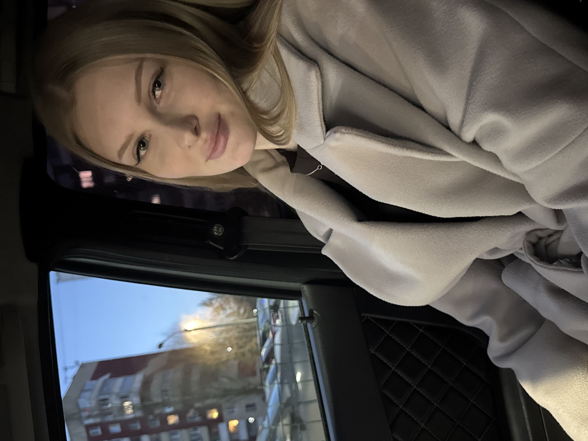 A young, light-skinned woman with blonde hair is in the back seat of a car. She is wearing a light gray coat or jacket. A building is visible through the window. The buildings are of standard modern construction, and appear to be located in Perm, Russia. The photo appears to have been taken at dusk. The car's interior is dark, with diamond stitching pattern on the door panel.