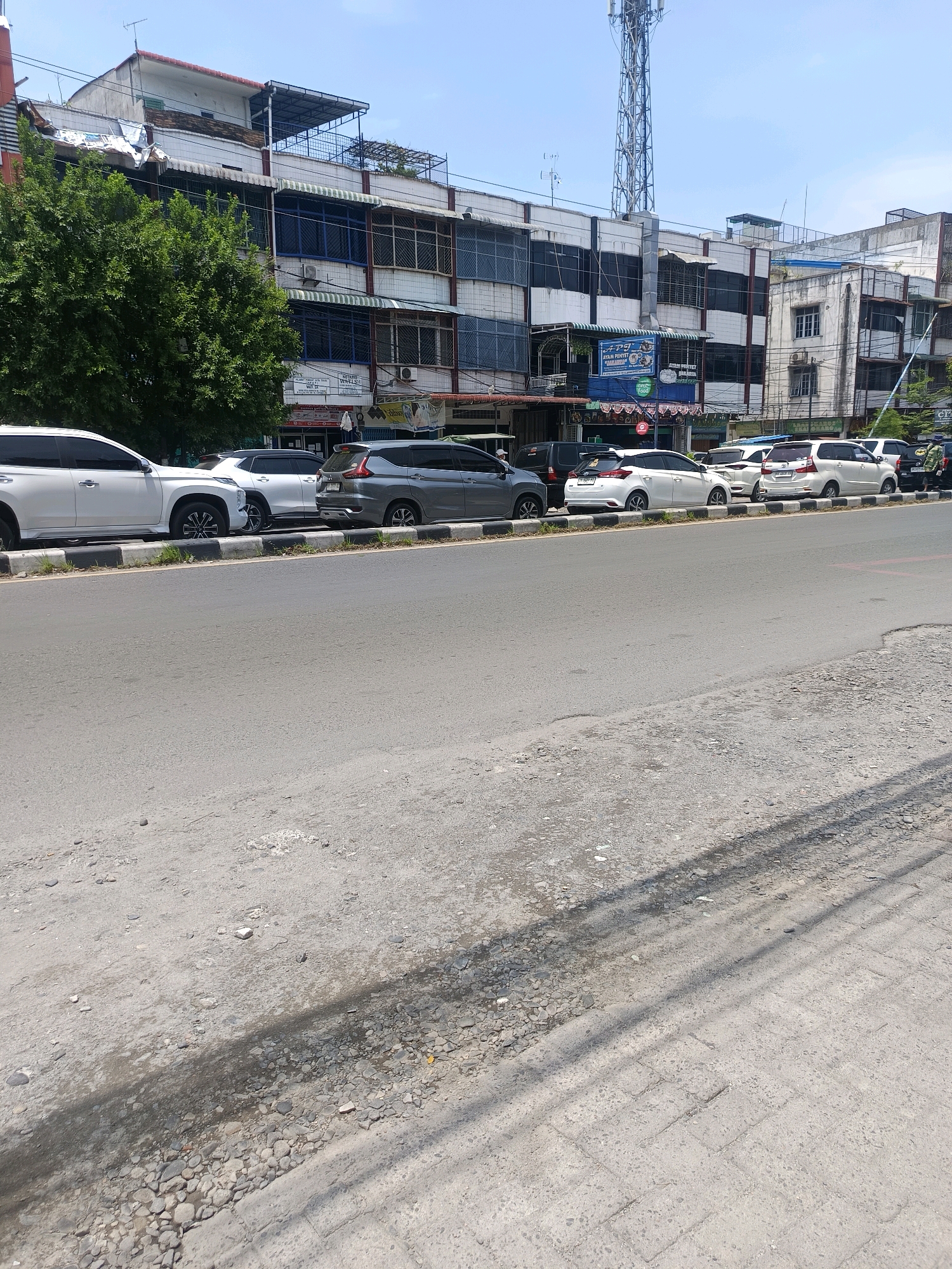 The image depicts a street scene in Percut Sei Tuan, Indonesia, under a clear blue sky. A row of parked cars lines the right side of the asphalt road, positioned next to a sidewalk constructed with alternating black and white painted curb stones. The buildings on the left side of the street are multi-story structures, some appearing commercial with awnings over windows and signage visible. A large, leafy green tree is situated on the far left. In the background, a tall telecommunications tower rises above the buildings. The road surface itself is rough and weathered in the foreground.