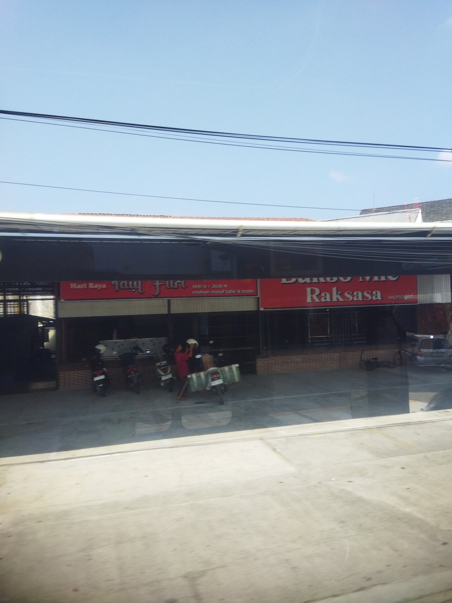 The image captures a street-level view of a commercial establishment in Wonokerto, Indonesia, under a clear blue sky. In the foreground, a reflective surface, likely a window or a car windshield, distorts the scene, creating a mirror effect of the concrete ground. Beyond this reflection, the establishment displays two prominent red signs. The sign on the left reads "Hari Raya" followed by stylized text that appears to be "Jamu" or a similar phonetic spelling, with additional Indonesian text beneath. The sign on the right is larger and features the text "Bakso Mie" followed by "Raksasa" in bold white lettering, and "wonogiri" in smaller text to its right.
In front of the establishment, several motorcycles are parked. The storefront itself appears to be open, with visible dark interiors. Two individuals are present near the entrance. One person, wearing a red shirt, is standing and appears to be engaged in some activity, possibly with objects on the ground. Another person, partially visible, is near the motorcycles. The building's facade is constructed with brickwork at the lower level. The overall impression is that of a bustling local business, likely a food establishment, in an Indonesian town.