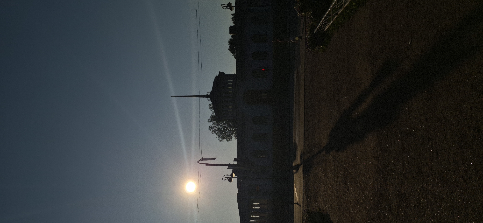 A vertical, low-angle shot captures a sun-drenched architectural scene at dusk. The sun, a bright orb in the upper left quadrant, casts a warm glow on the horizon, with its rays emanating outwards and creating a visible light streak across the hazy, twilight sky. A silhouetted building, featuring a domed structure topped with a spire and surrounded by a low-lying, colonnaded facade, dominates the mid-ground. A solitary streetlamp with two arms stands in the foreground, its silhouette stark against the sky. The ground in the foreground is dark, textured, and appears to be a park or pedestrian area, with the long, stretched shadows of what appears to be a person and a lamppost extending across it. A traffic light with a red illuminated bulb is visible on the building's facade, indicating a pedestrian crossing in the mid-ground, where a person is walking. The environment suggests an urban setting during the golden hour.