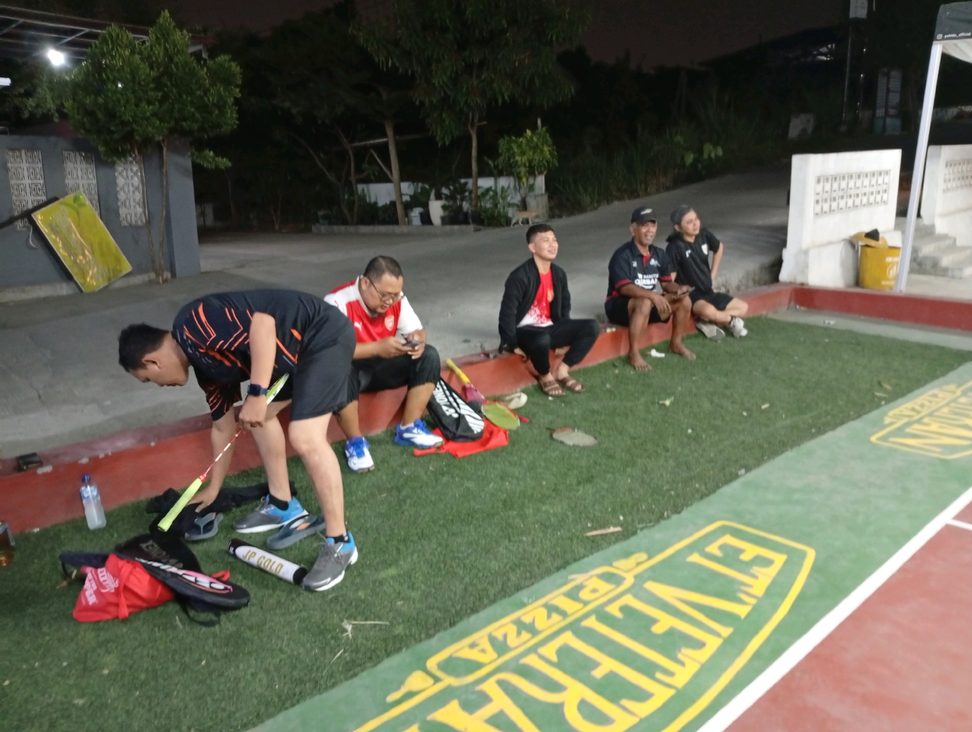The image depicts a group of men gathered on a green, artificial turf field in what appears to be an outdoor recreational area in Caringin, Indonesia. The setting is dimly lit, suggesting it is evening or nighttime. In the foreground, a man in an orange and black shirt is bending over, appearing to pick up a badminton racket from a red bag and black case on the ground. Several other men are seated on a low wall or ledge behind him, some looking towards the camera and others looking away. They are dressed in casual athletic wear, including t-shirts, shorts, and one man is wearing a black cap. Badminton rackets, a water bottle, and a white cylindrical tube labeled "JP GOLA" are visible on the ground near the man in the foreground. The background features trees, a building with a patterned facade, and a portion of a white structure. The ground in front of the seated men is lined with green turf, and beyond that, a red surface with green sections painted with yellow lettering is visible, part of a court marking. The overall atmosphere suggests a break during or after a sports activity.