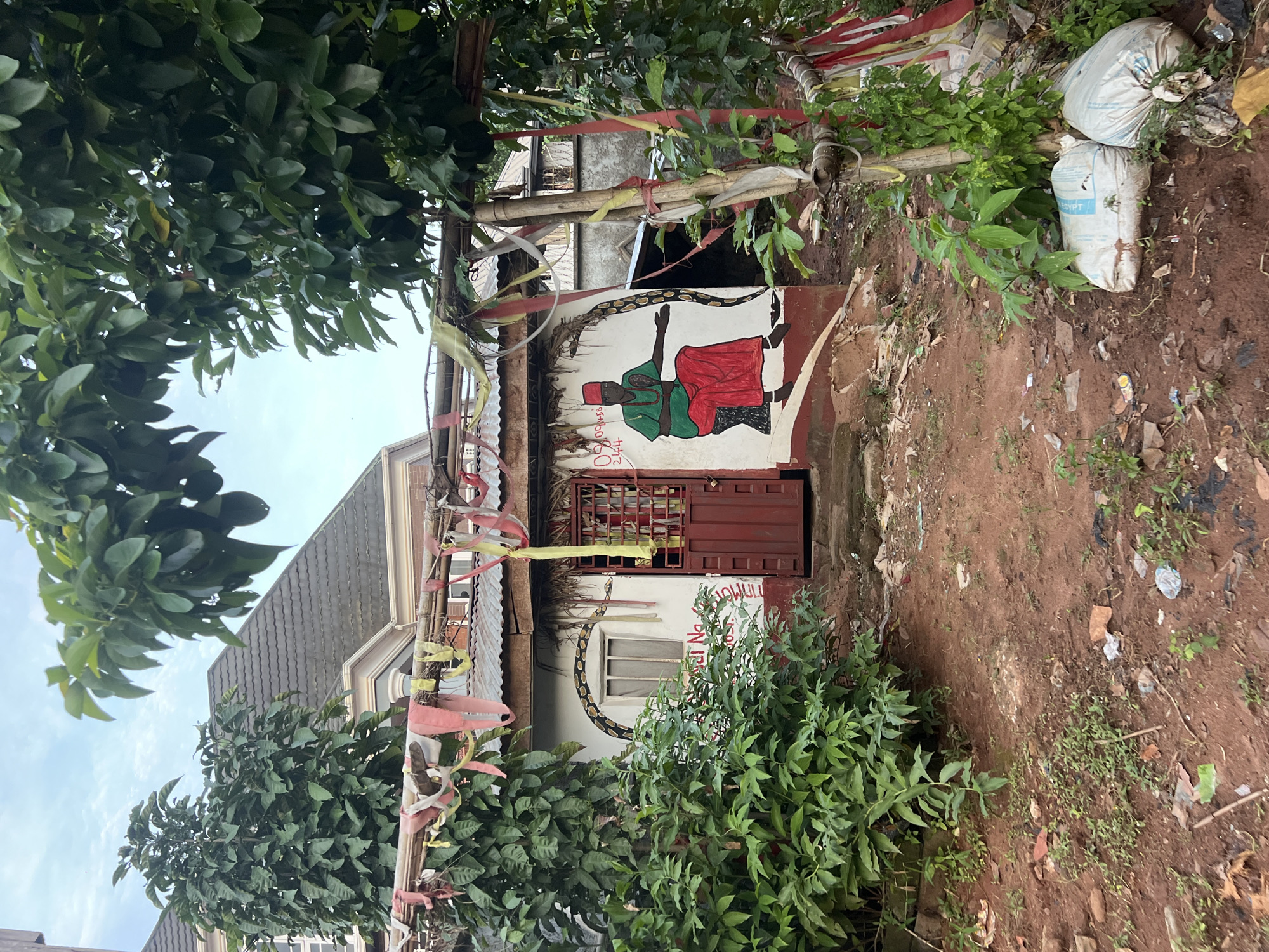 In Nkpor, Nigeria, a modest, aged dwelling is captured in a slightly elevated outdoor shot. The building, with its corrugated metal roof and white painted walls, exhibits a rustic charm. The facade is adorned with a vibrant mural depicting a seated figure in a red skirt and green shirt, a red cap, and a beaded necklace. A painted snake arches over a window. Colorful streamers, red, yellow, and white, are strung across a bamboo-like structure in front of the building. Lush green foliage frames the scene, with trees and bushes flanking the dwelling. The ground in front of the house is unpaved, red earth, littered with debris, including plastic bags and fragments of what appear to be discarded building materials. Two large, white sacks, likely filled with goods or refuse, are placed near the entrance, partially obscured by vegetation. The sky above is overcast. The overall impression is one of a lived-in, perhaps spiritual, space, set against a backdrop of everyday Nigerian life.