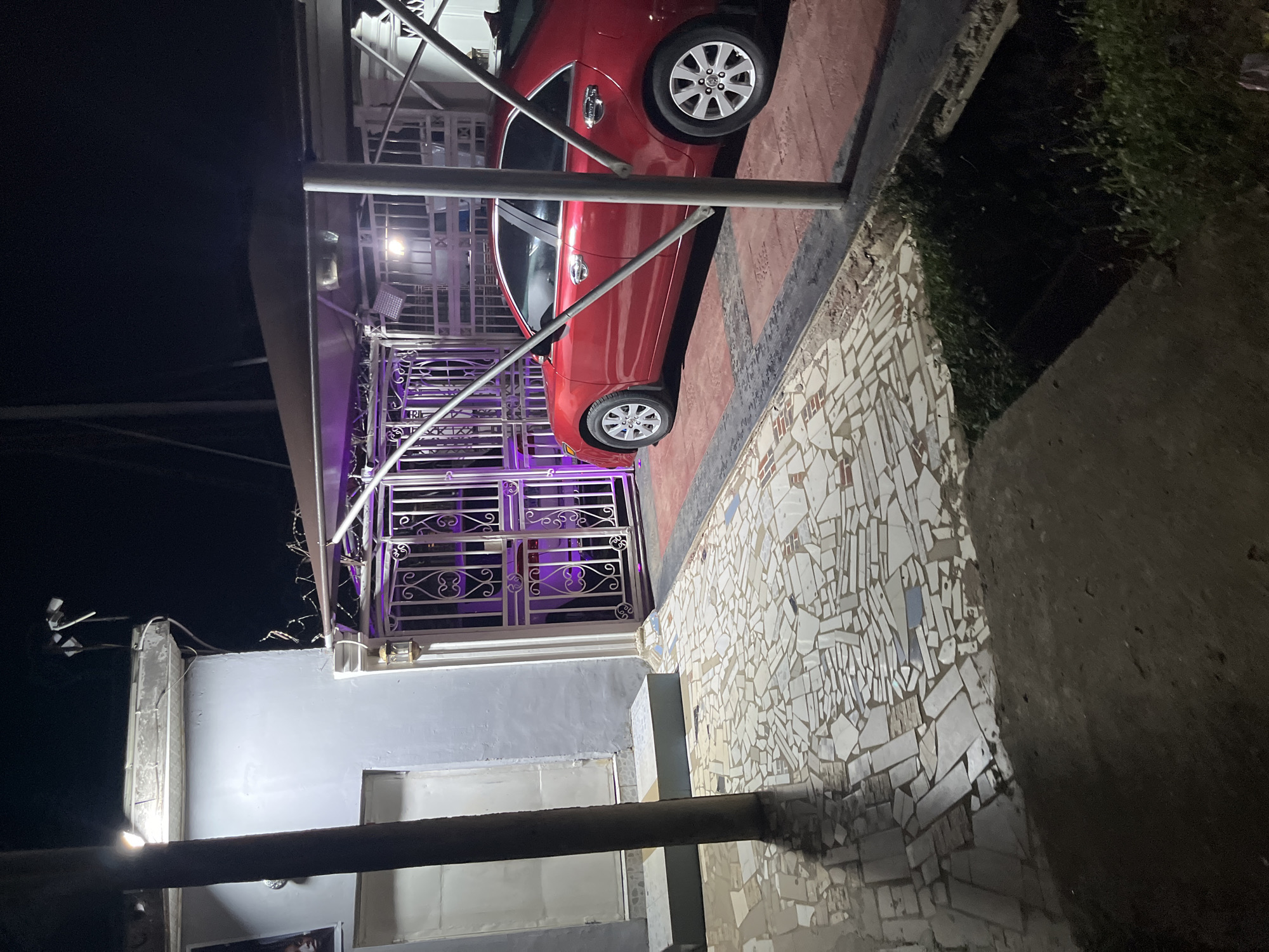 The image depicts a night scene at a residence in Abuja, Nigeria. A red sedan is parked under a carport with a corrugated metal roof, supported by metal poles. The carport is illuminated by overhead lights. In front of the carport is a white metal gate with ornate scrollwork, which is lit with purple lights, revealing a glimpse of another vehicle behind it. Adjacent to the gate is a light-colored building with a white door. The ground in front of the gate and leading to the carport is paved with a mosaic of irregular white and blue tiles. To the left, a dark pole stands vertically, casting a shadow. A pathway with two rectangular concrete steps is visible to the left of the gate. The overall environment suggests a residential property with security features, including the gated entrance and outdoor lighting.